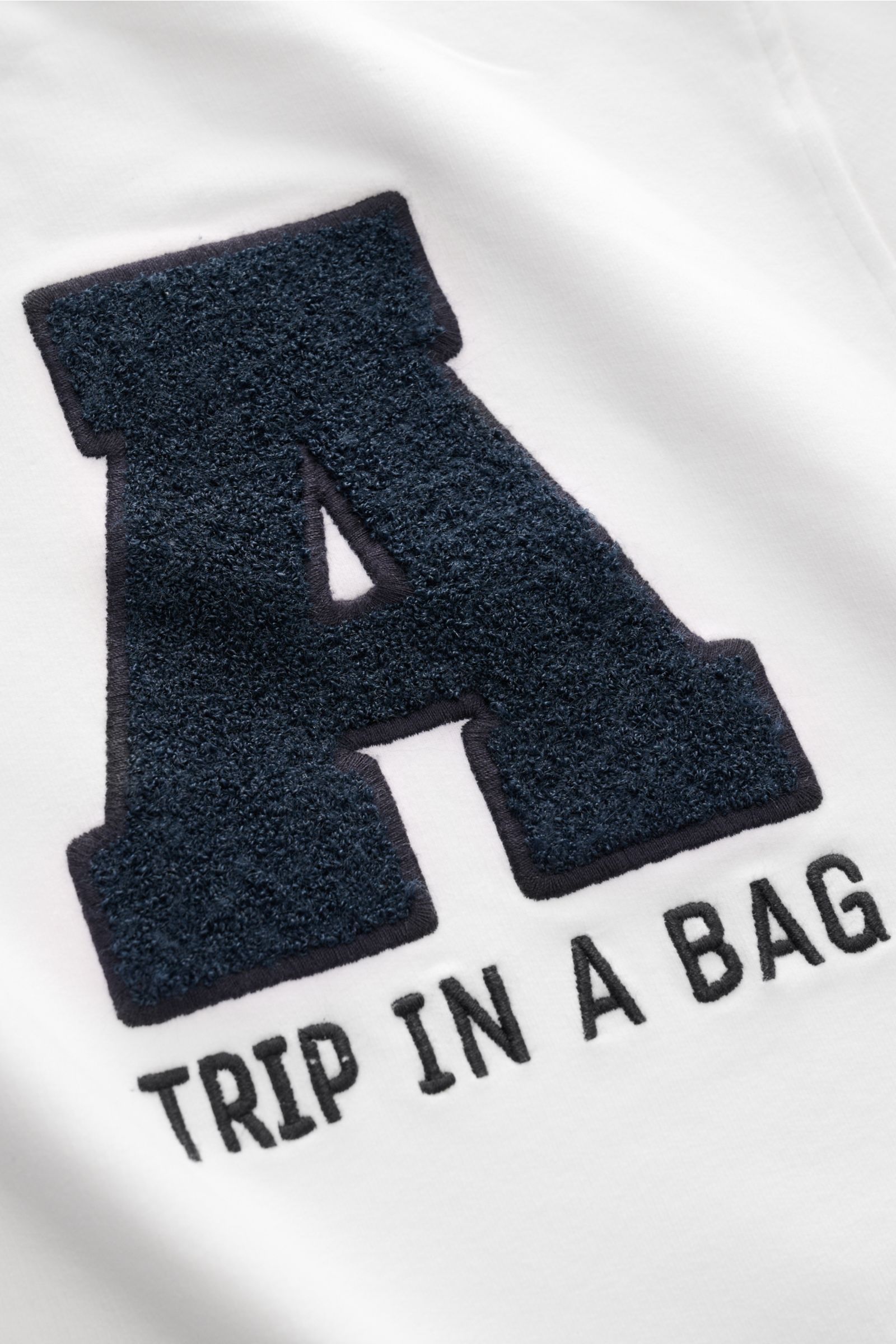 Close-up front view of the offwhite 04651/ A trip in a bag Kapuzenpullover 'IF Hoodie' showing a textured navy blue letter 'A' and 'TRIP IN A BAG' text on soft brushed cotton jersey fabric.