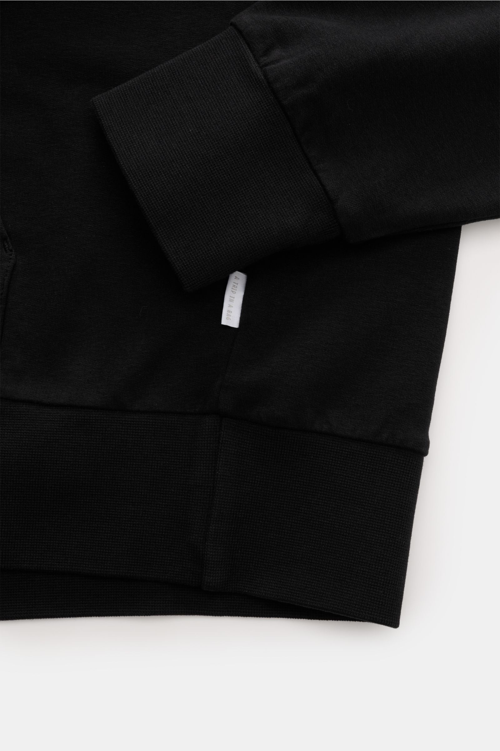 Close-up view of the black 04651/ A trip in a bag Hooded jumper 'IF Hoodie' showing the ribbed cuff, waistband, and small woven side label in brushed stretchy cotton with a soft feel.