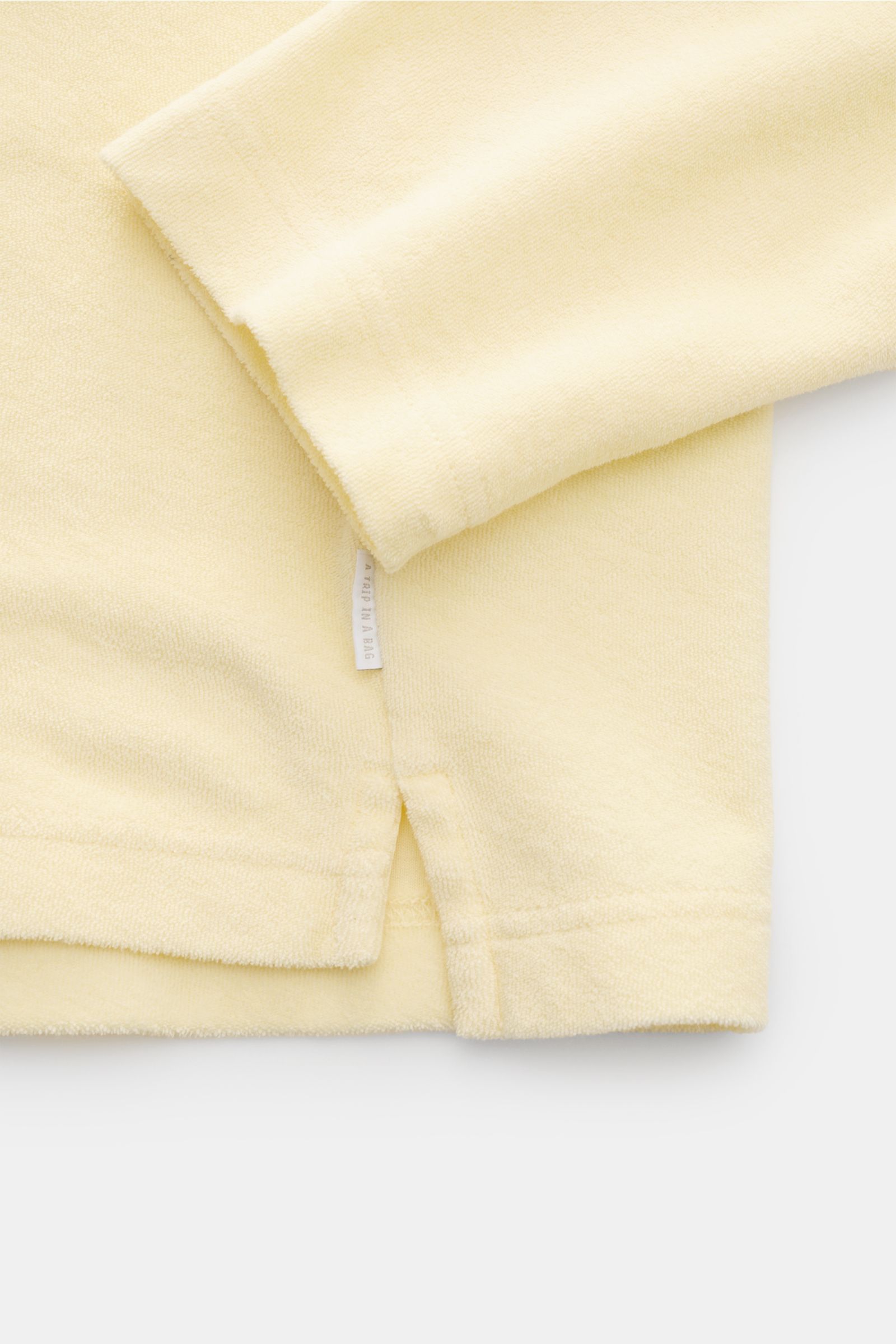 Nahaufnahme des pastellgelben 04651/ A trip in a bag Frottee-Kapuzenpullovers 'Terry' aus Baumwoll-Frottee, Regular Fit, mit weichem Griff, Kapuze und kleinem Weblabel in der Seitennaht, fotografiert von der Seite.