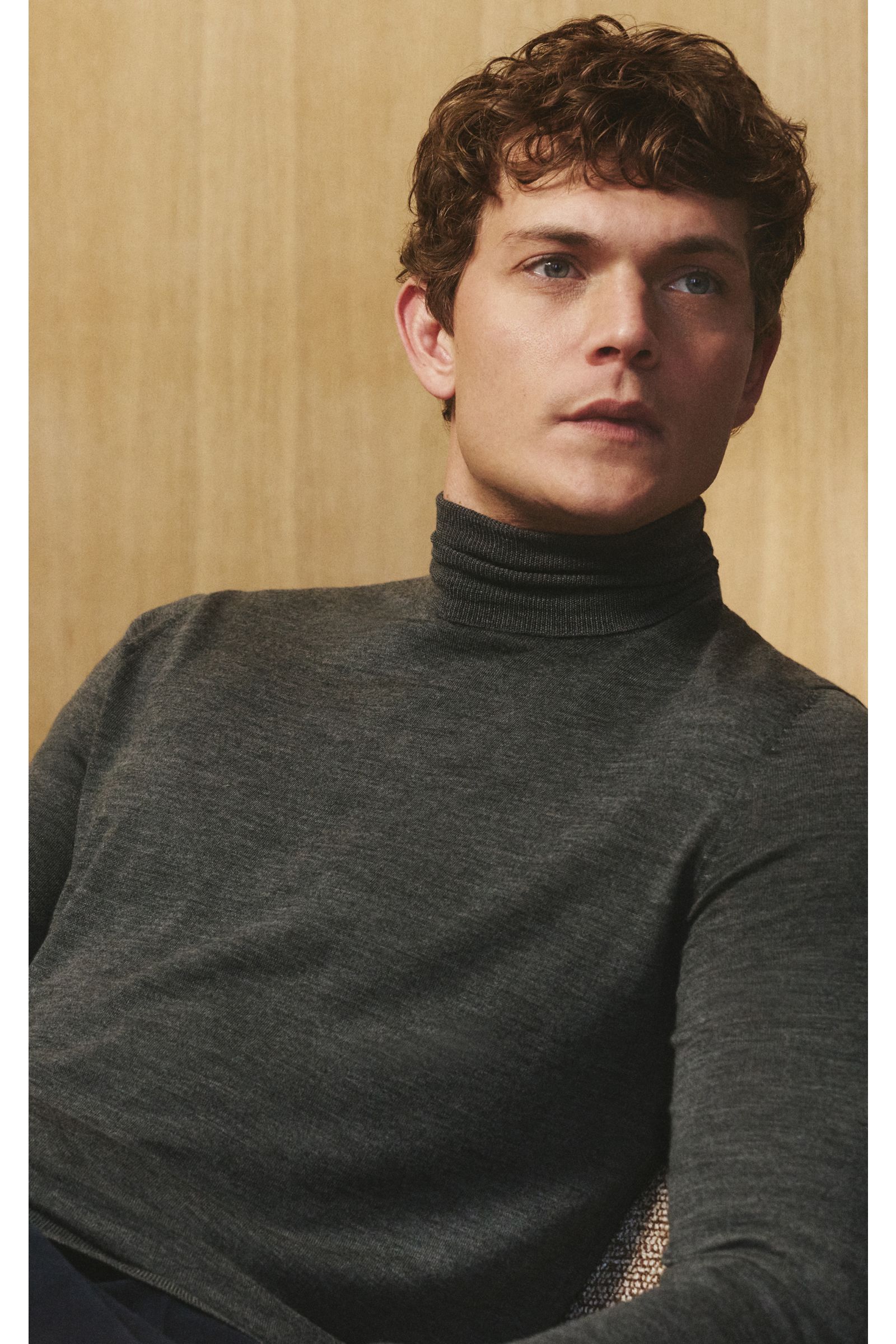 von Braun Merino Rollkragenpullover anthrazit meliert, superfein gestrickt aus reiner Merinowolle, Slim Fit mit anliegendem Rollkragen, flexible Rippbündchen, seitliche Nahaufnahme am Modell vor hellem Holz-Hintergrund.