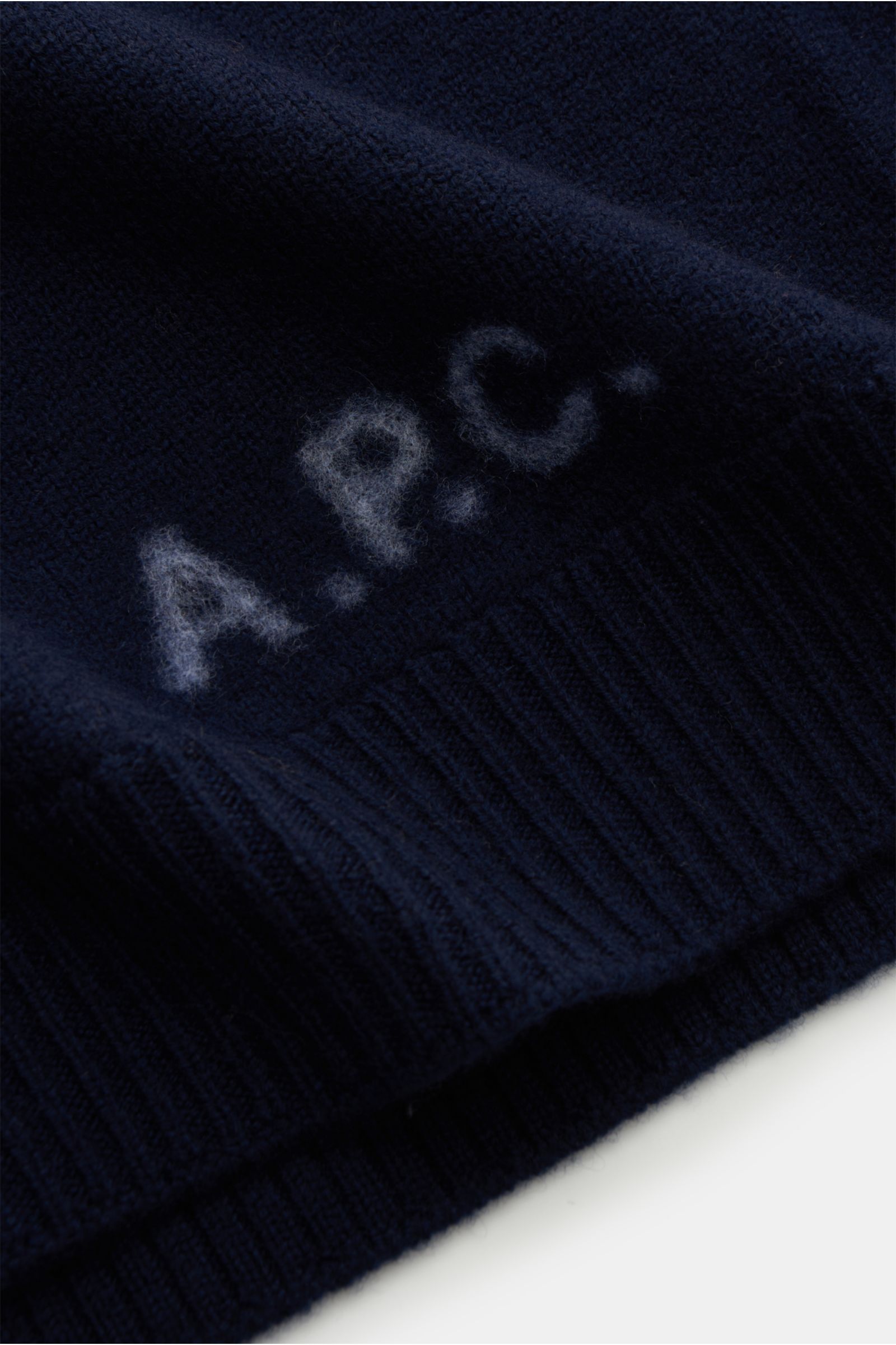 Close-up shot from the front-bottom perspective of A.P.C. Rundhalspullover 'Edward' navy, showcasing its reine Wolle, weicher Griff, mittelfeines Strickbild, Slim Fit, flexible Rippbündchen, and a dezentes Logo auf der Vorderseite.