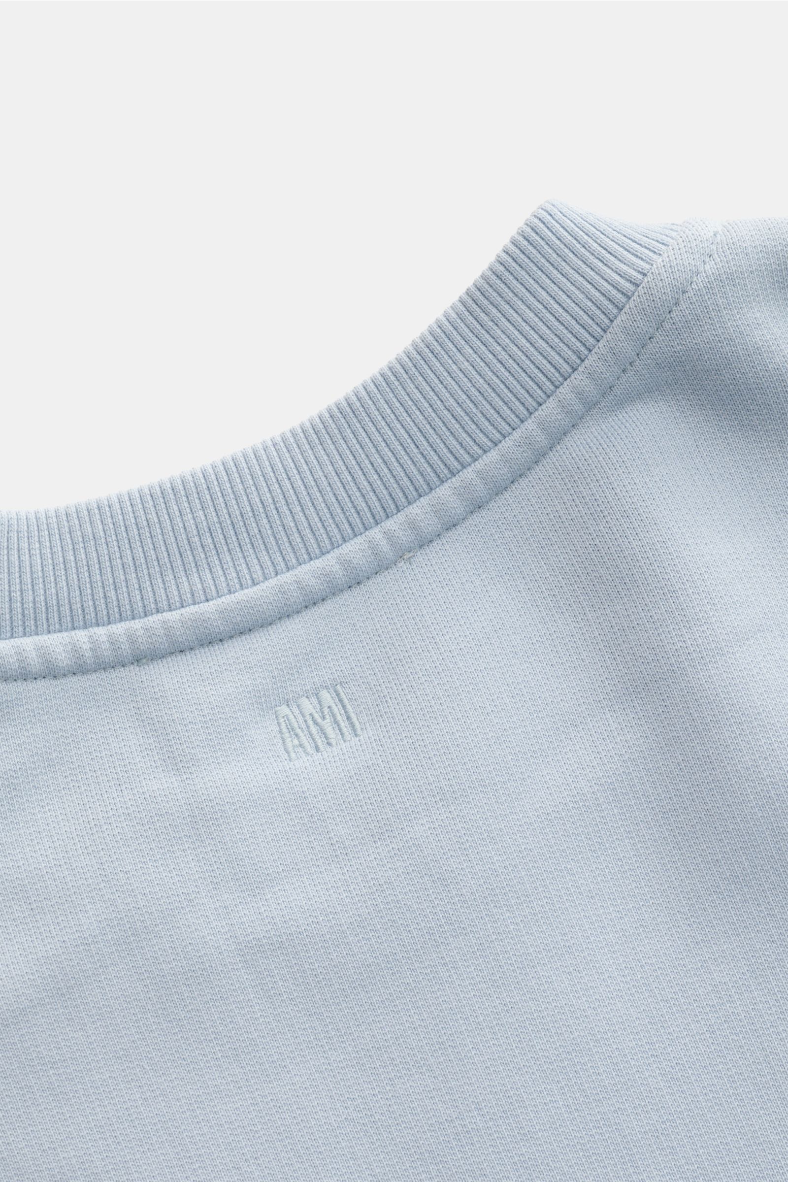 A close-up rear view of the Ami Paris Rundhals-Sweatshirt hellblau, showing light blue cotton fabric with ribbed round neckline, subtle AMI stitching on the back, and a soft, slightly textured surface. A subtle heart-stitching on the chest elevates this b
