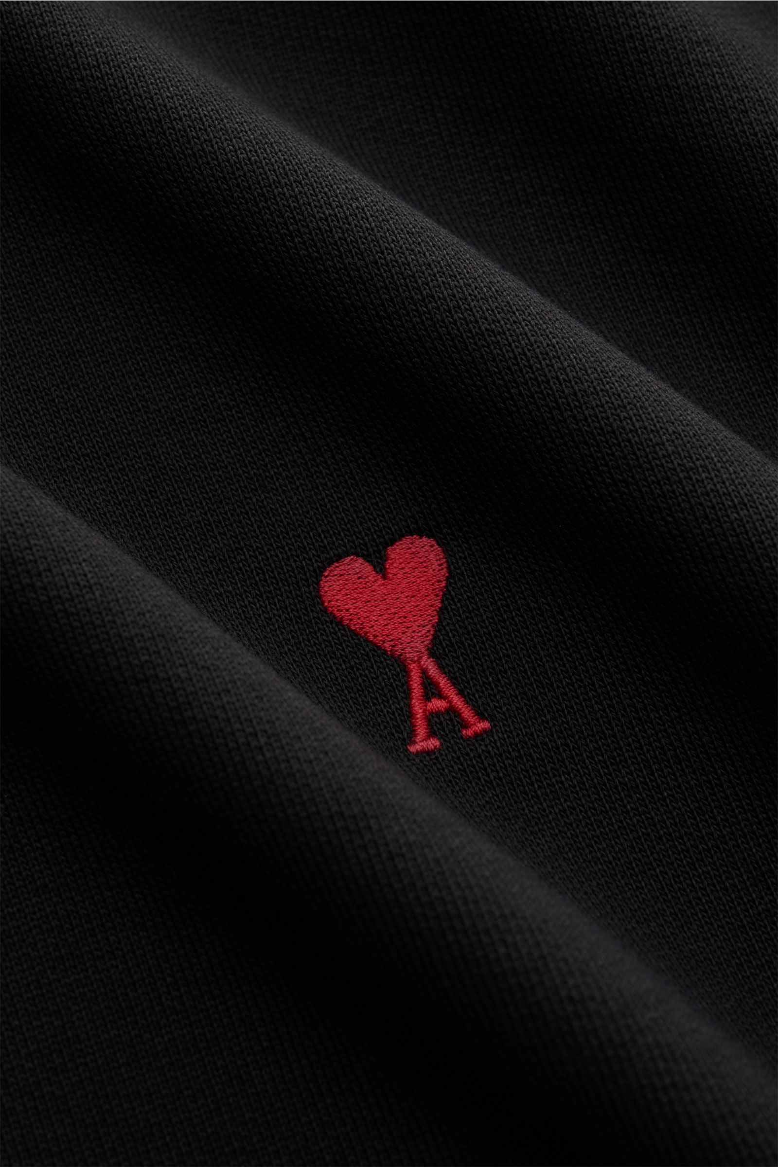 Close-up of the Ami Paris Rundhals-Sweatshirt schwarz fabric, showing the red heart-stitching with an 'A' on black organic cotton from a top-down perspective. Love is in the air! Das markentypische Herz-Stitching auf der Brust wertet dieses Basic-Sweatshi