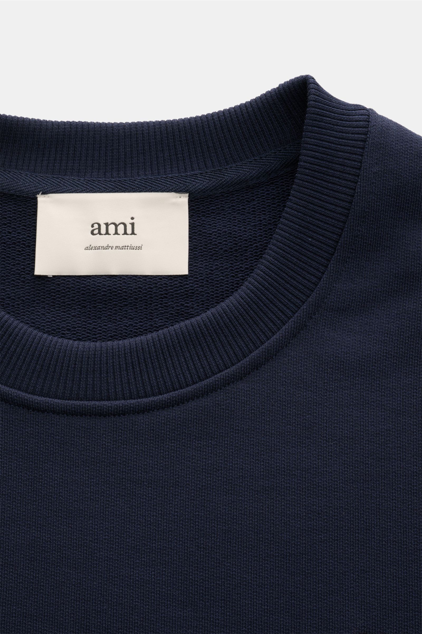 A close-up front view of the dark blue Ami Paris Rundhals-Sweatshirt dunkelblau showing the ribbed round neckline and the 'ami' label inside the collar. Love is in the air! Das markentypische Herz-Stitching auf der Brust wertet dieses Basic-Sweatshirt aus