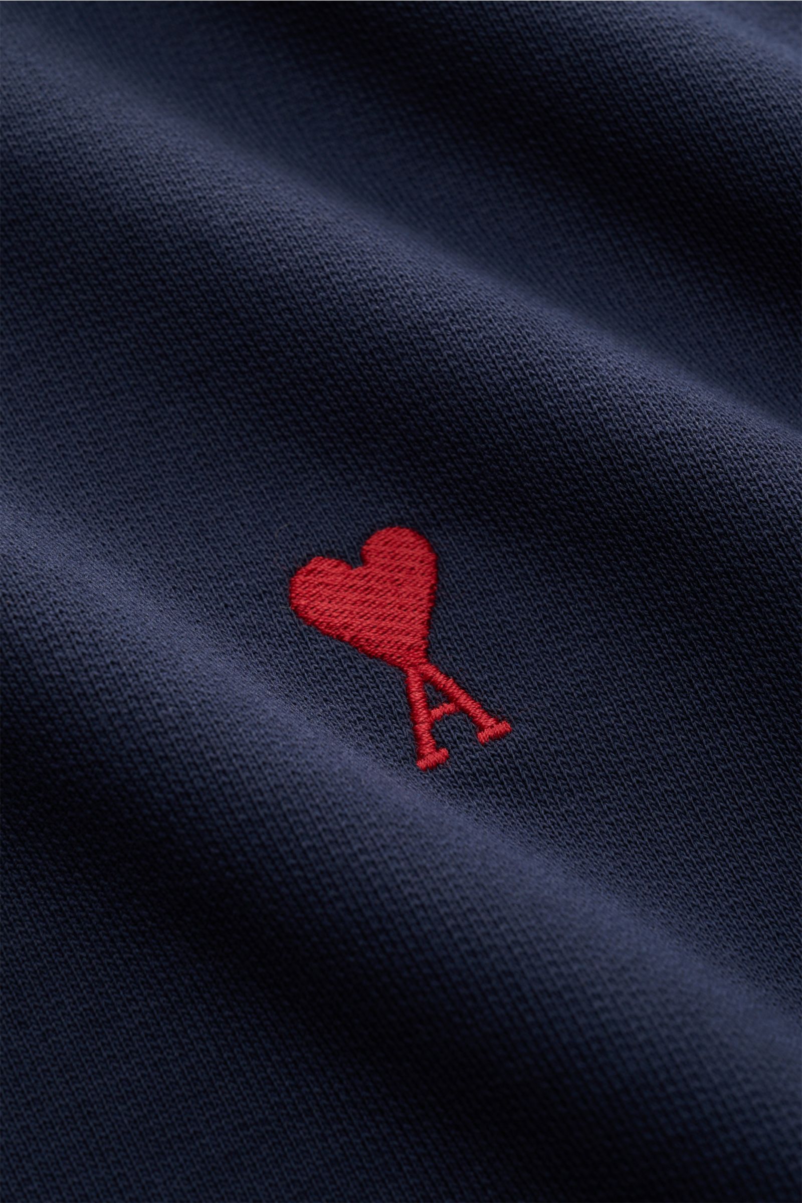 Close-up photo of the Ami Paris Rundhals-Sweatshirt dunkelblau showing the red heart-stitching with an 'A' on dark blue organic cotton fabric, highlighting its texture and details from a front perspective. Love is in the air! Das markentypische Herz-St