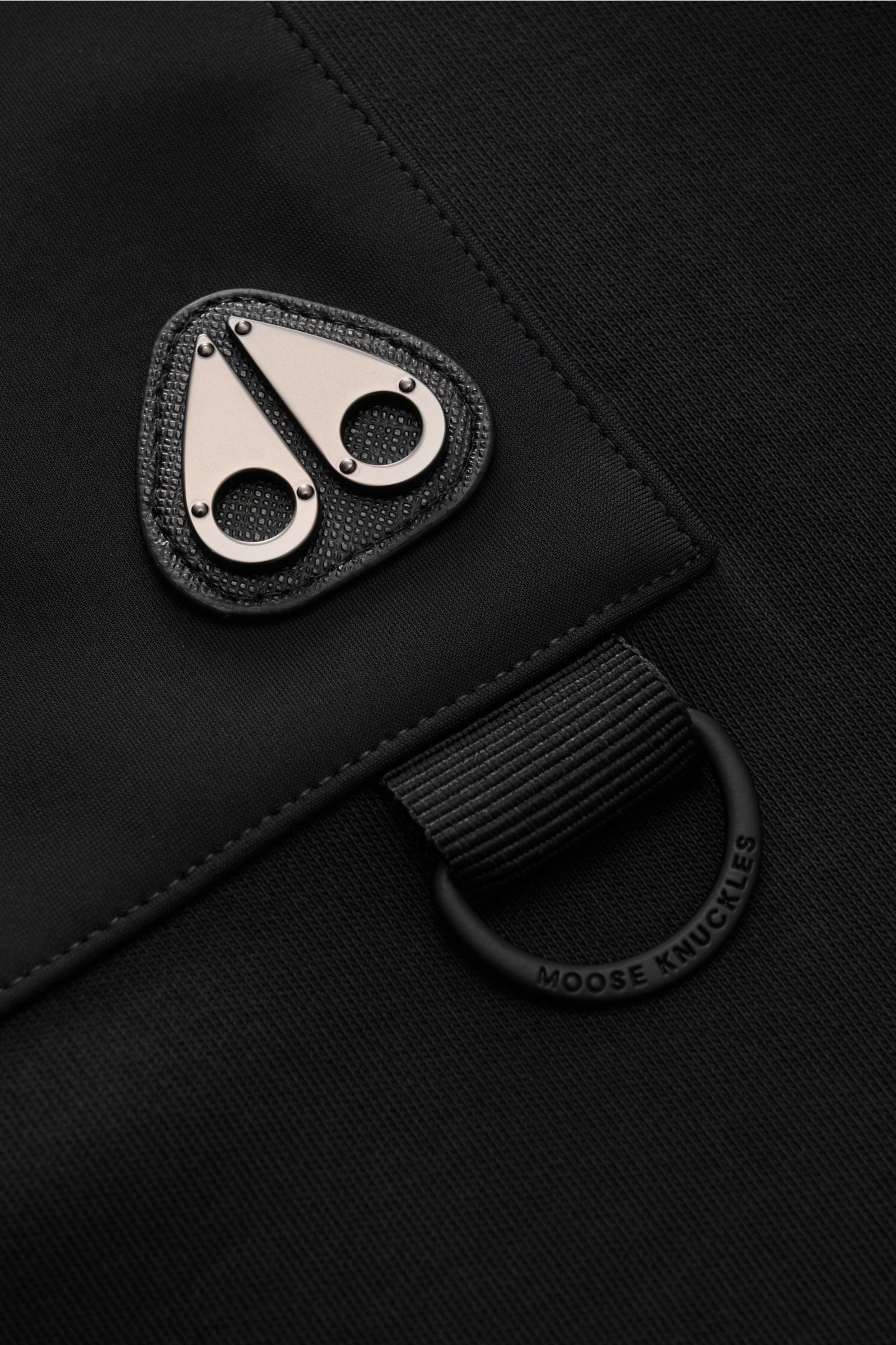 Close-up front view of the zipped breast pocket with metal Moose Knuckles logo on the black Moose Knuckles Crew neck sweatshirt 'Dalon Crew'. 

Refined sportswear: A practical zip pocket featuring the signature MOOSE KNUCKLES logo in metal stands out o