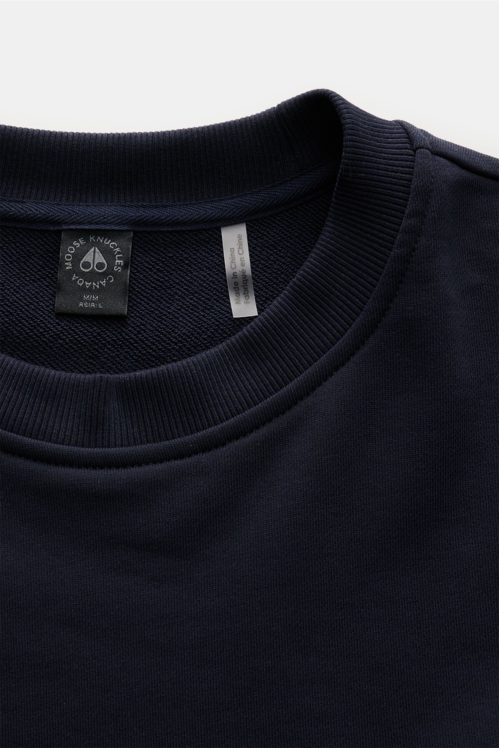 Nahaufnahme des Moose Knuckles Sweatshirts 'Hartsfield Crew' navy mit Rundhalsausschnitt, weichem Griff und Metall-Logo-Patch am Oberarm, fotografiert von oben.