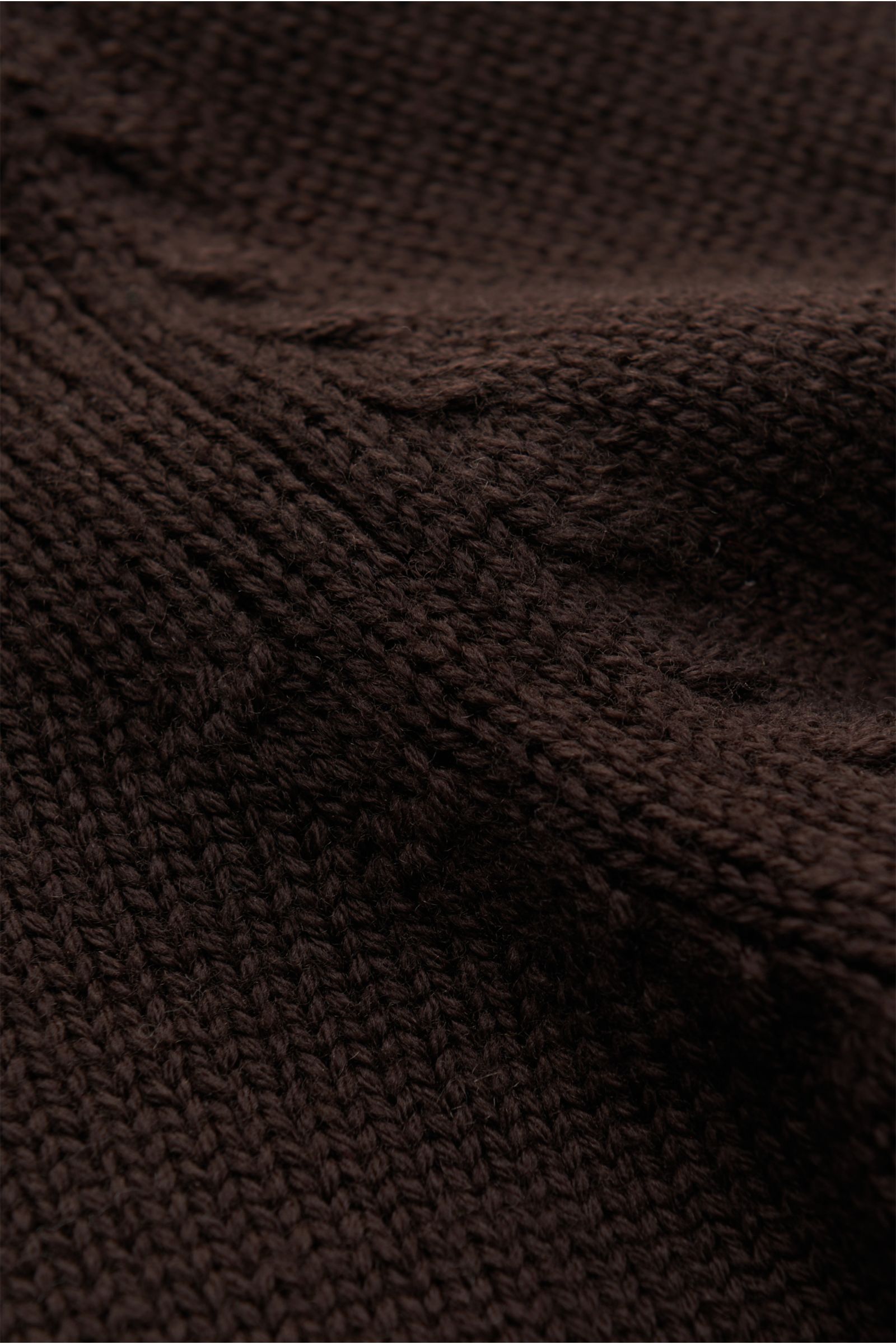 Nahaufnahme des dunkelbraunen B.F. Knitwear Merino Rundhalspullovers aus reiner Merinowolle mit weichem Griff, mittelgrobem Strickbild, Slim Fit, Rundhalsausschnitt und flexiblen Rippstrick-Bündchen, fotografiert aus schräger Perspektive.