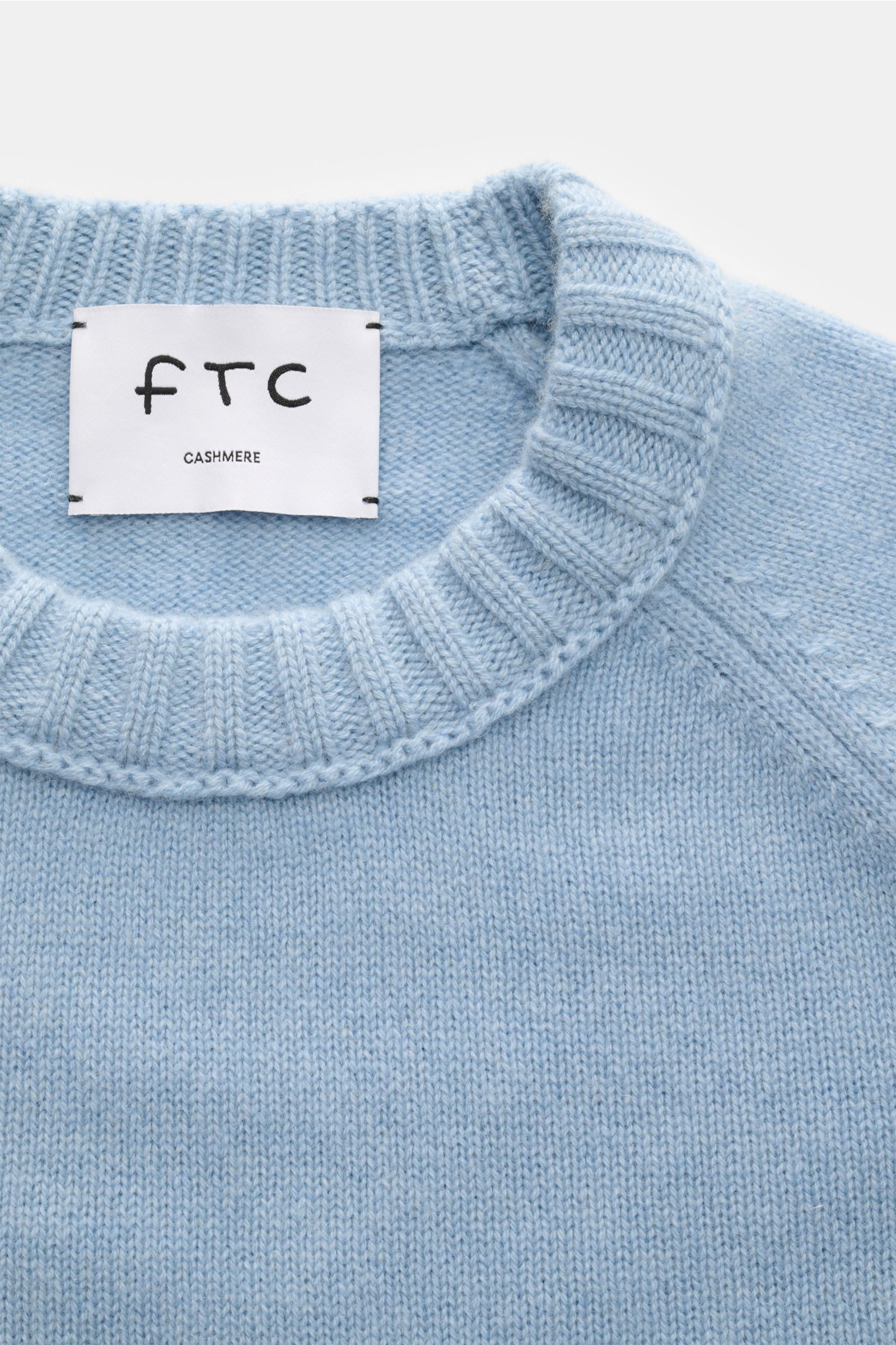 Close-up top-down view of FTC Cashmere crew neck jumper light blue, showing medium-fine knit, slim fit, crew neck, raglan sleeves, ribbed bands; pure fair trade cashmere, OEKO-TEX® certified, very soft feel.