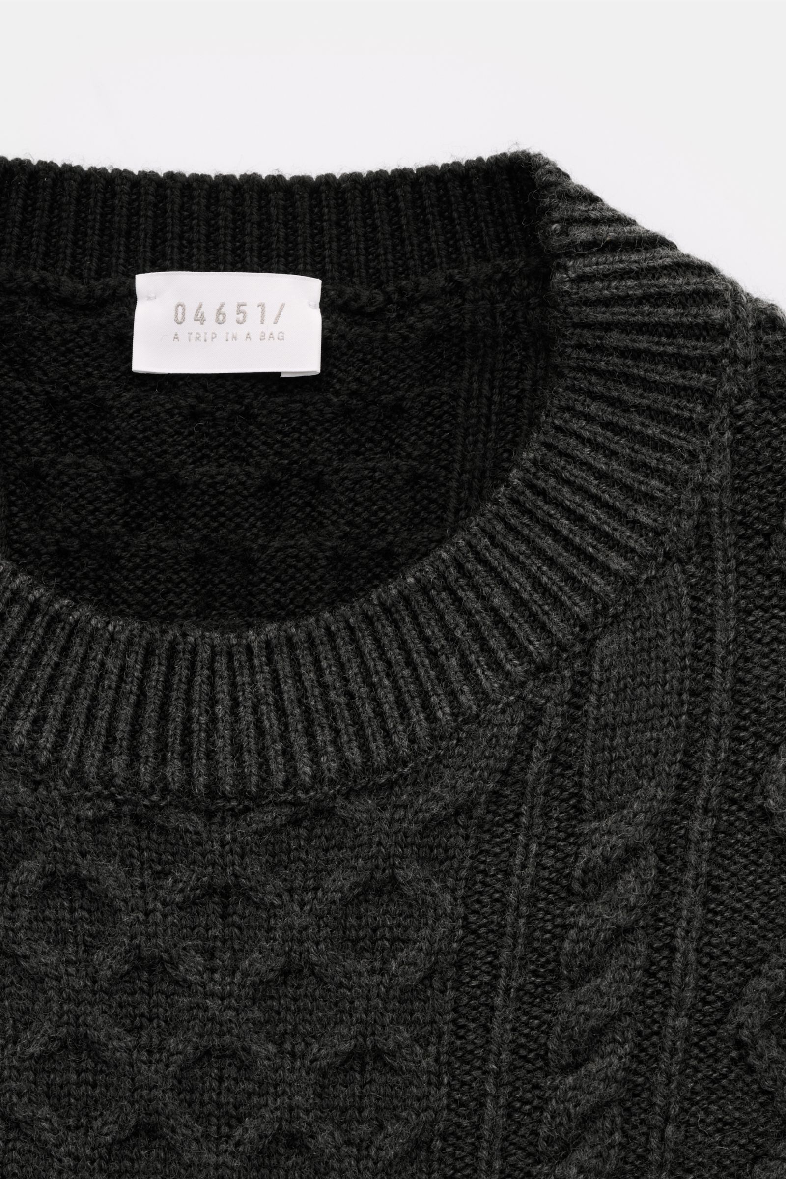 Close-up top-down view of the 04651/ A trip in a bag Rundhalspullover 'Foggy Cable Crew' dunkelgrau, showing a classic cable knit pattern, ribbed crew neckline, and soft wool texture. Klassischer Zopfstrick neu interpretiert: Der Rundhalspullover von 0465