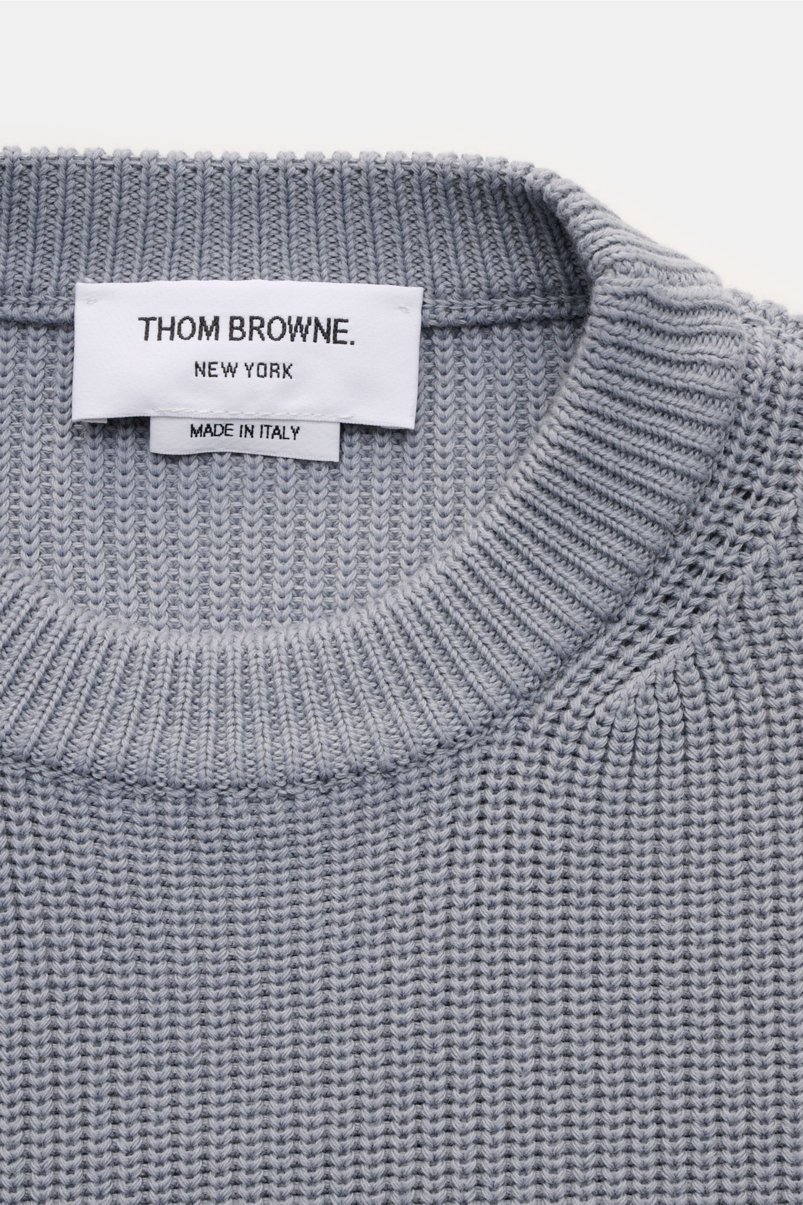 Close-up top-down view of Thom Browne Rundhalspullover rauchblau showing fine medium knit, round neckline, and brand label. Stilvoll durch die kalte Saison: Der Rundhalspullover aus reiner Baumwolle von THOM BROWNE setzt mit seinen charakteristischen Sign