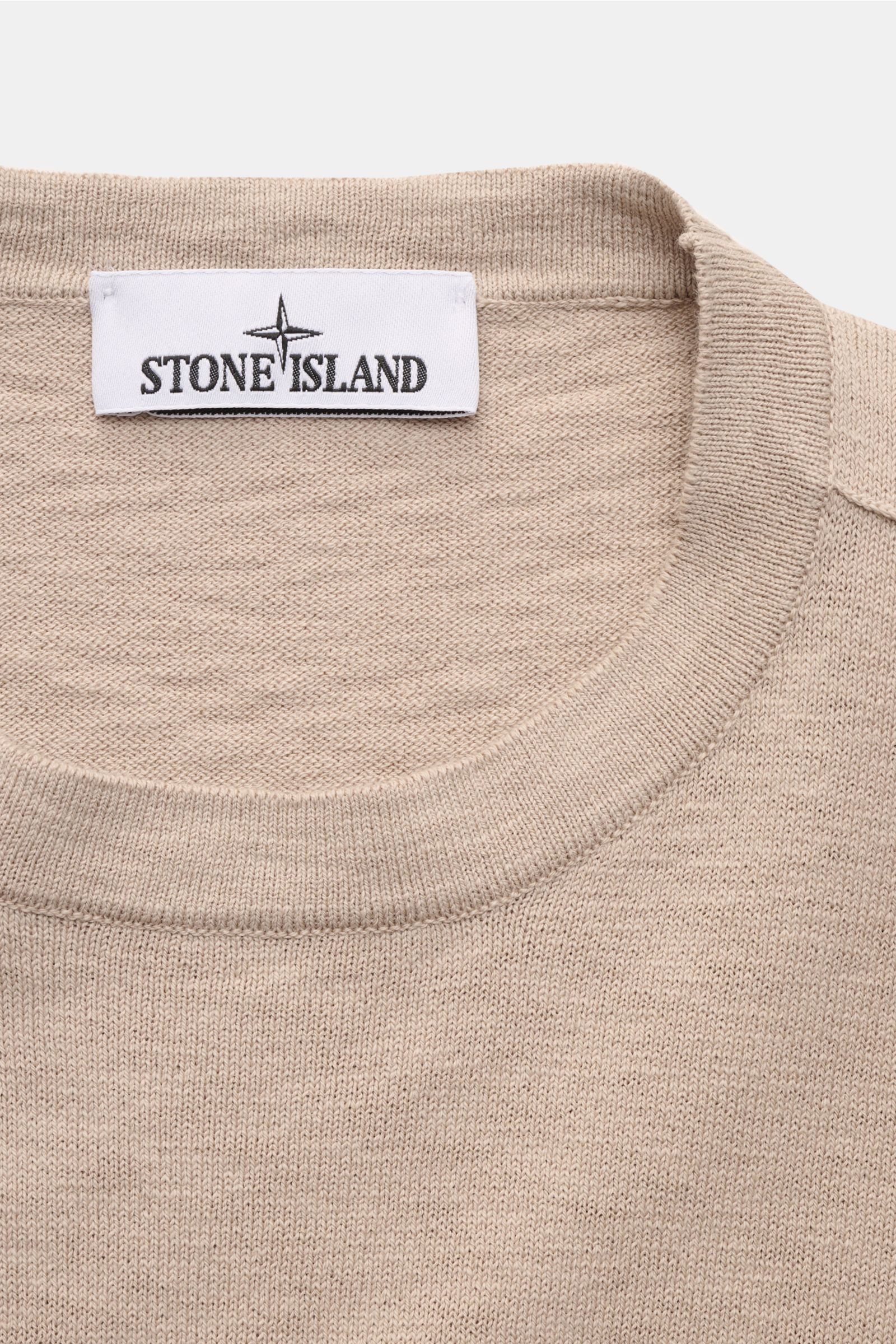 Nahaufnahme des Stone Island Rundhalspullover beige von oben, mittelfeines Strickbild, weicher Griff, Slim Fit, Rundhalsausschnitt, Kompass-Badge am Oberarm.