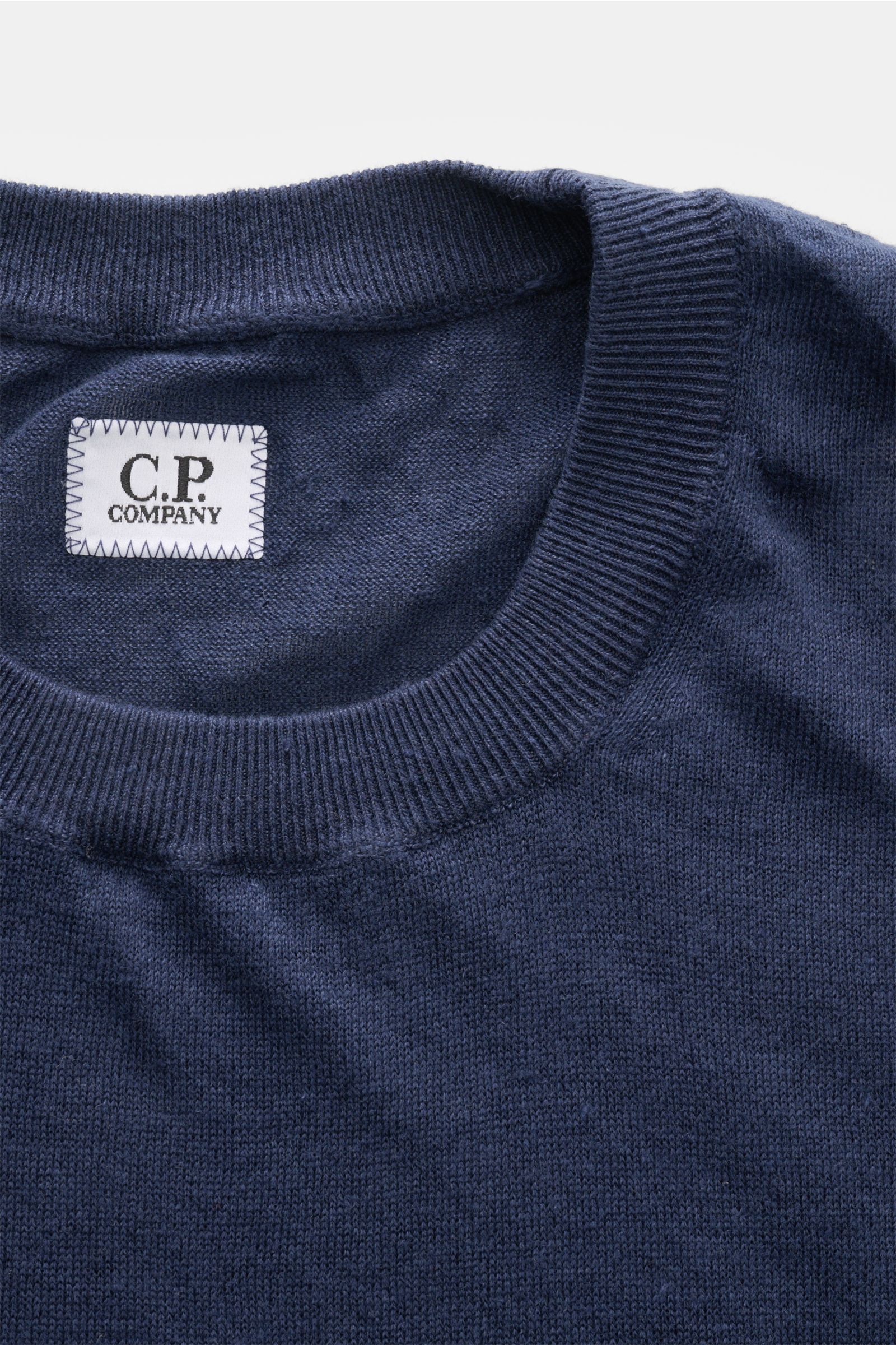 Nahaufnahme des C.P. Company Kurzarmpullovers navy aus feinstem Leinenstrick, weicher Griff, Regular Fit, elastische Rippbündchen und Weblabel am Saum, fotografiert von oben.