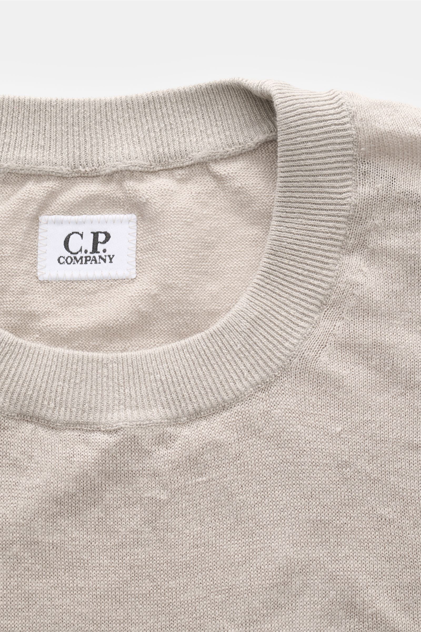 C.P. Company Kurzarmpullover grau, Nahaufnahme des Rundhalsausschnitts mit feinem Leinenstrick, elastischen Rippbündchen und kleinem Weblabel am Saum, Regular Fit, weicher Griff, feines Strickbild, fotografiert von oben.