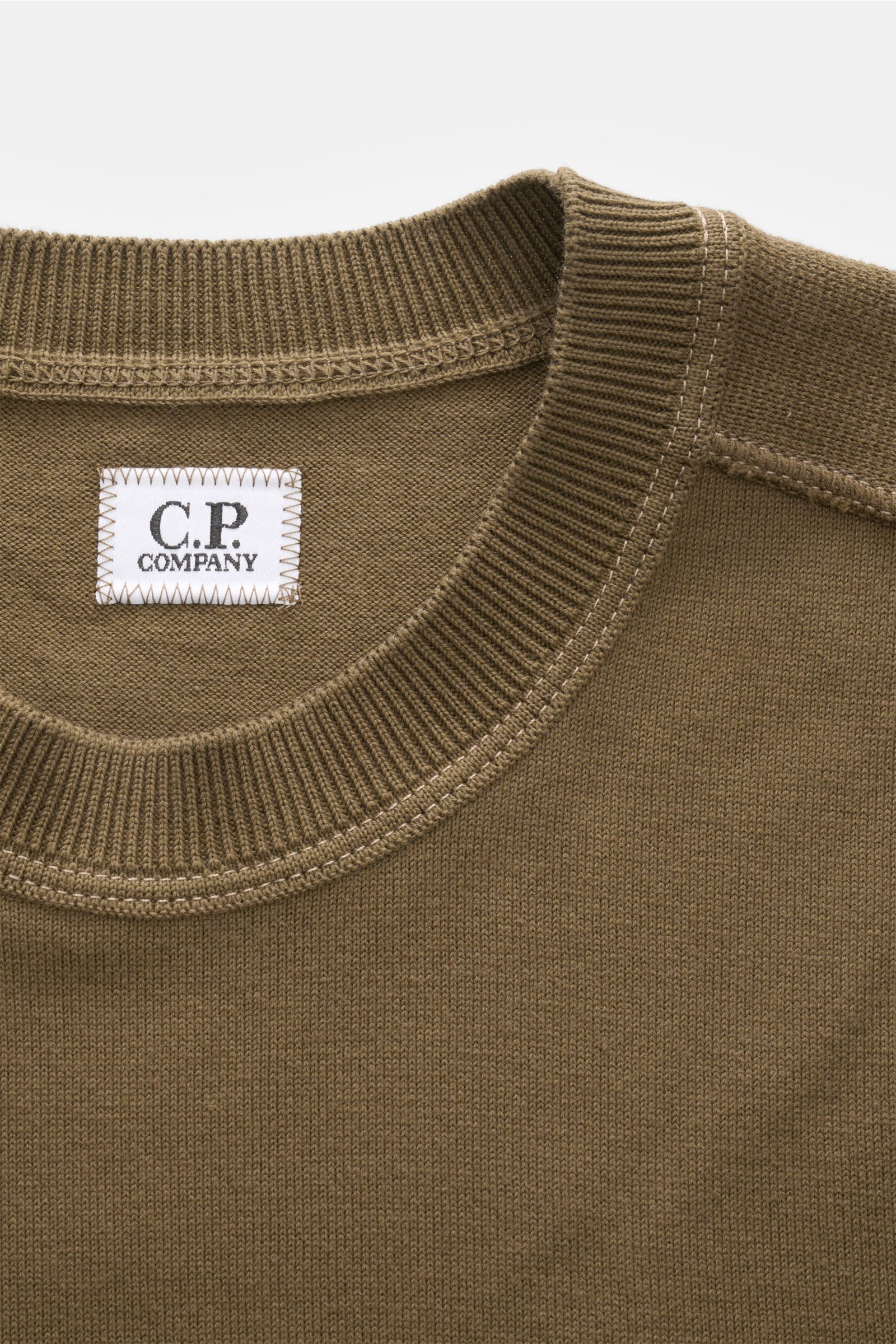 Nahaufnahme des olivfarbenen C.P. Company Rundhalspullover, fotografiert aus schräger Draufsicht, mit feinem Strick und Label innen.