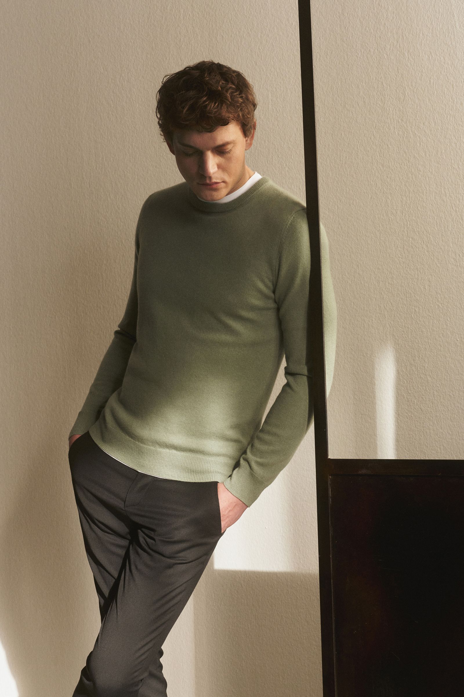 Junger Mann lehnt seitlich an Wand, trägt von Braun Cashmere Rundhalspullover salbei mit Slim Fit, Rundhals, weichem Griff, feinem Strickbild.