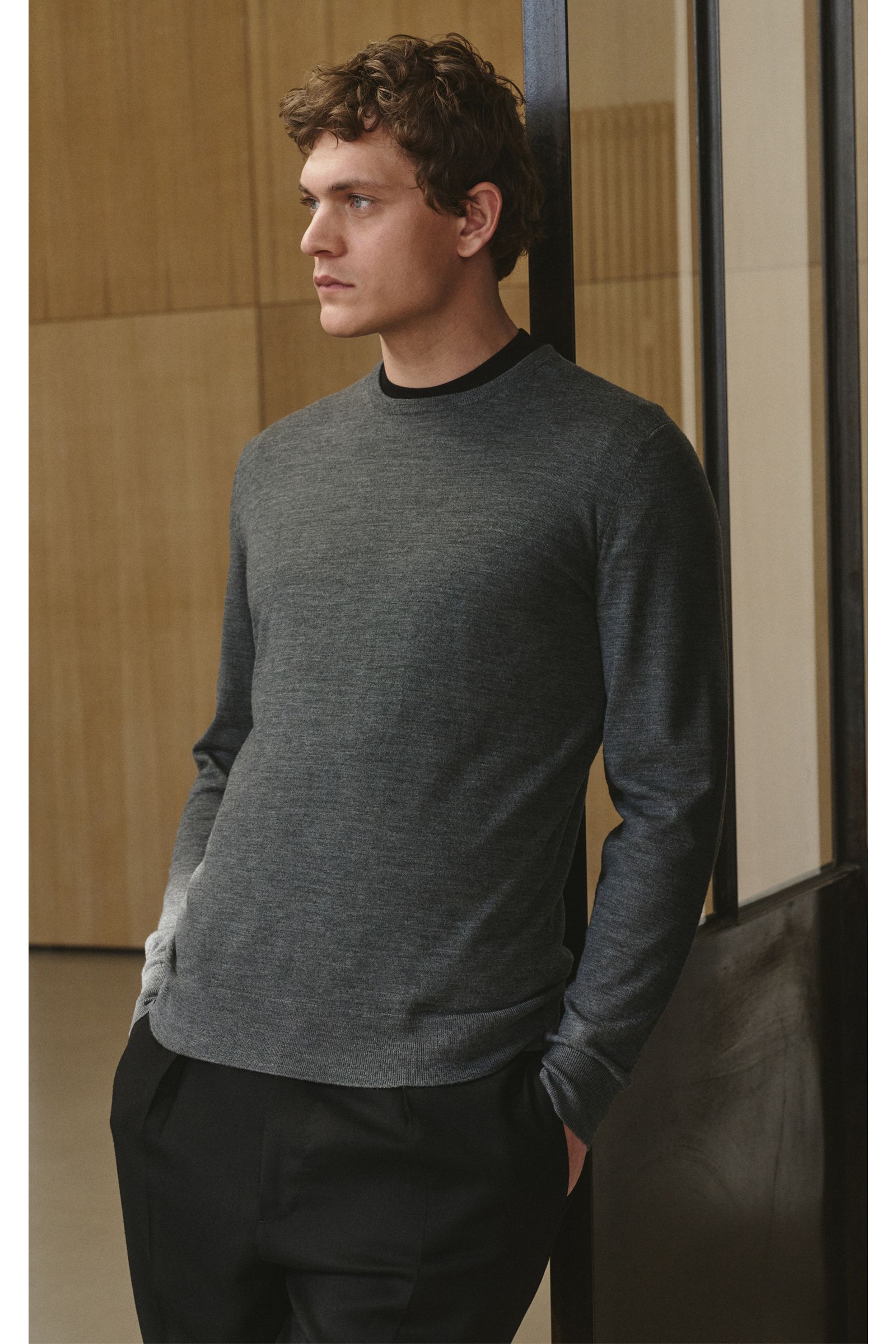 Mann von vorne links im Profil, trägt von Braun Merino Rundhalspullover anthrazit meliert aus reiner Merinowolle, Slim Fit, Rundhalsausschnitt, feines Strickbild, weicher Griff, elastische Rippbündchen, klassisches Basic für die Übergangszeit.