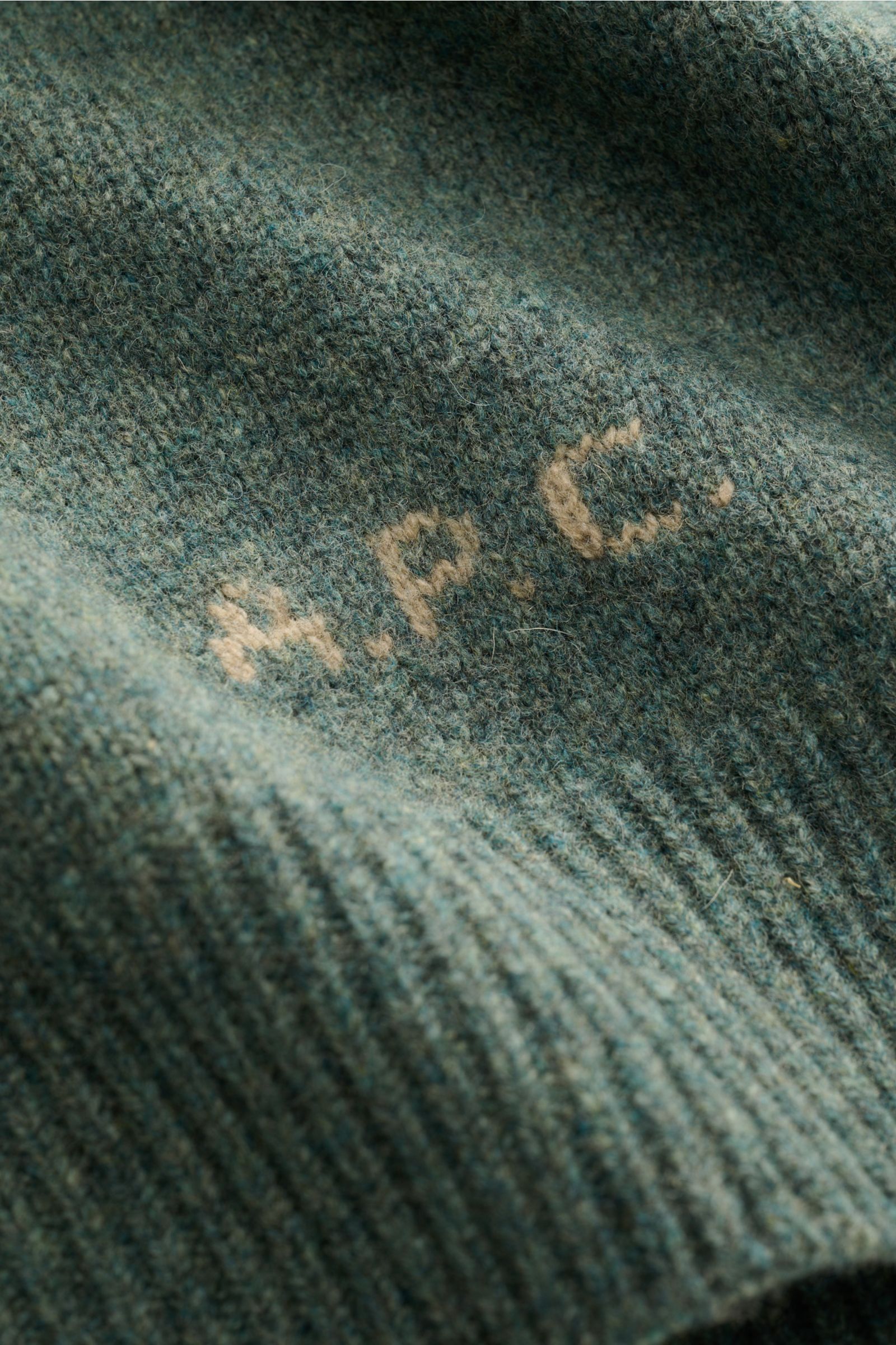 Nahaufnahme des A.P.C. Rundhalspullover 'Edmond' graugrün meliert aus reiner Wolle mit kontrastfarbenem Logo am Bund, Detailansicht.