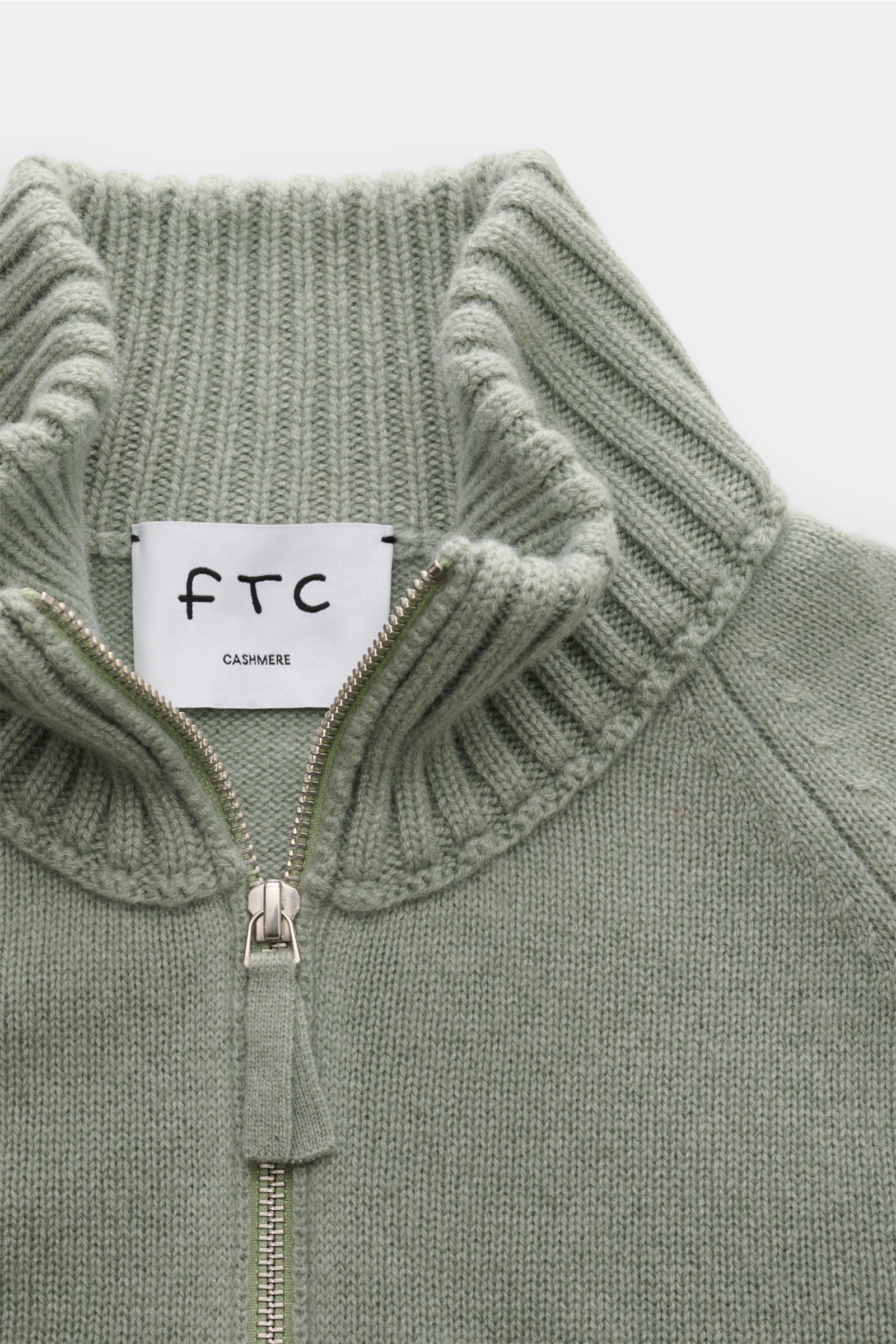 Nahaufnahme des FTC Cashmere Troyer graugrün von oben, mit angesetztem Troyer-Kragen, Zipper, Raglanärmel und Rippbündchen aus weichem Fair-Trade-Cashmere.