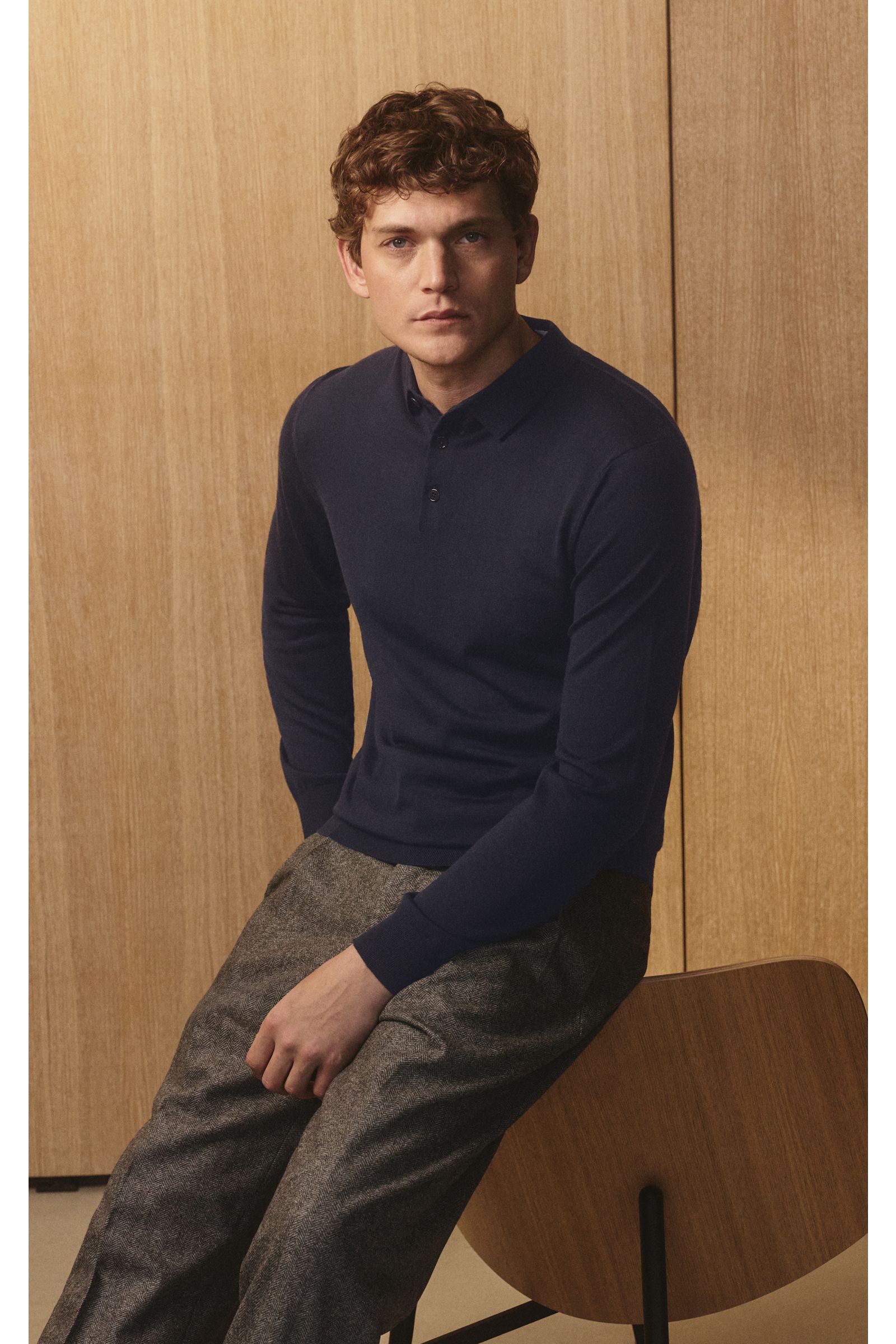 Mann sitzt seitlich auf Stuhl, trägt von Braun Merino Feinstrickpolo navy aus reiner Merinowolle, Slim Fit, schmaler Polokragen, glatte Knopfleiste, komfortable Rippbündchen.