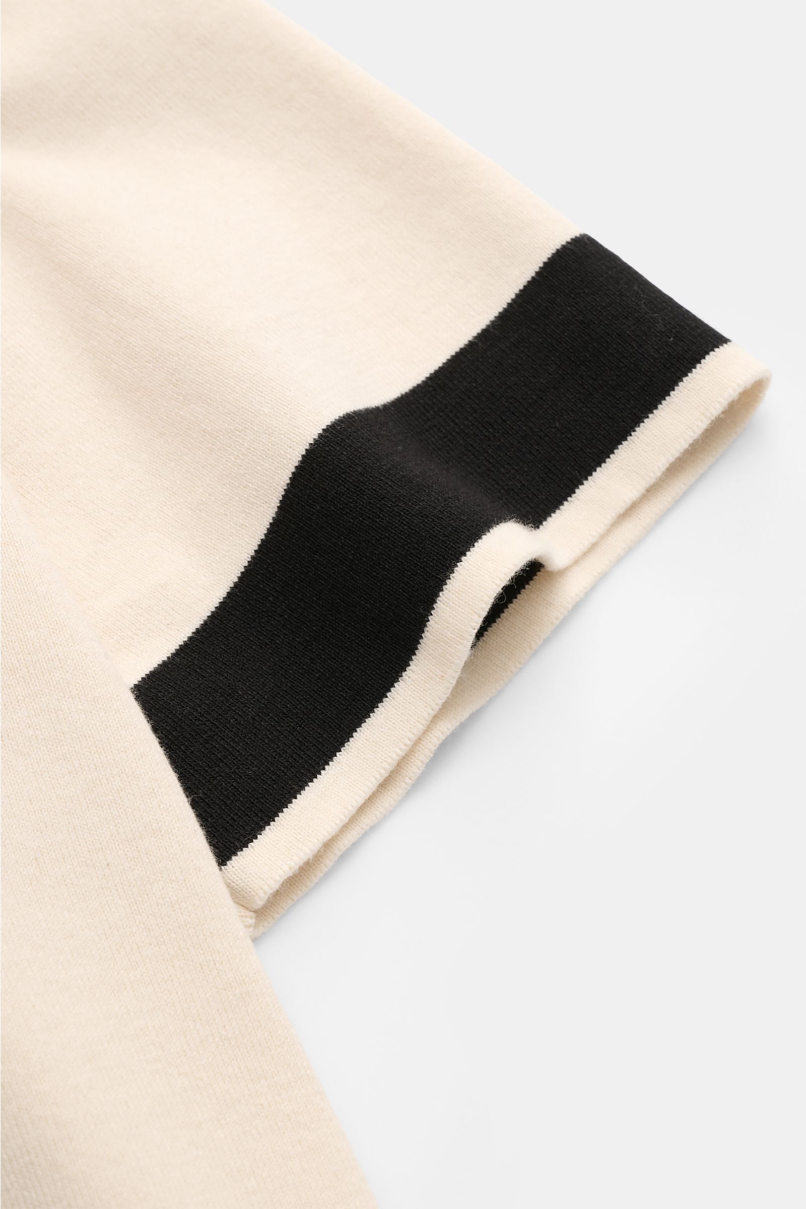 Close-up of the sleeve hem of the 04651/ A trip in a bag Short sleeve knit shirt 'Colour Block' cream/black, showing the fine knit texture and distinctive black colour block design, photographed from a side angle. 

An eye-catching piece for summer: Th