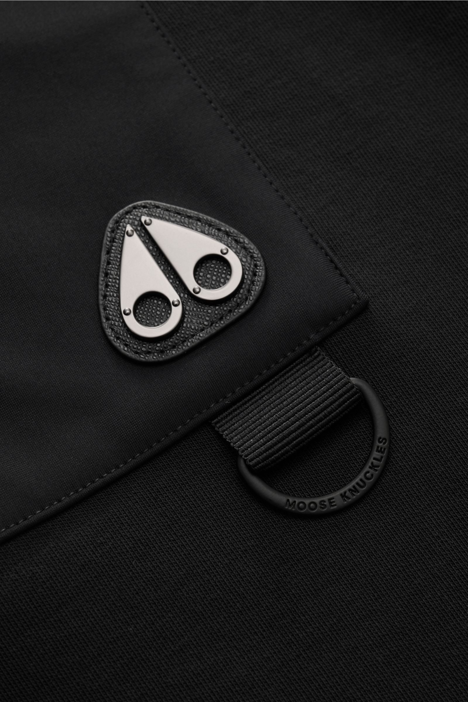 Close-up angled view of the Moose Knuckles Sweat-Troyer 'Dalon' schwarz showing the black fabric, metal logo on chest pocket, and branded elastic loop. Sportiv durch den Herbst: Der Sweat-Troyer von MOOSE KNUCKLES überzeugt als vielseitiges Basic für l