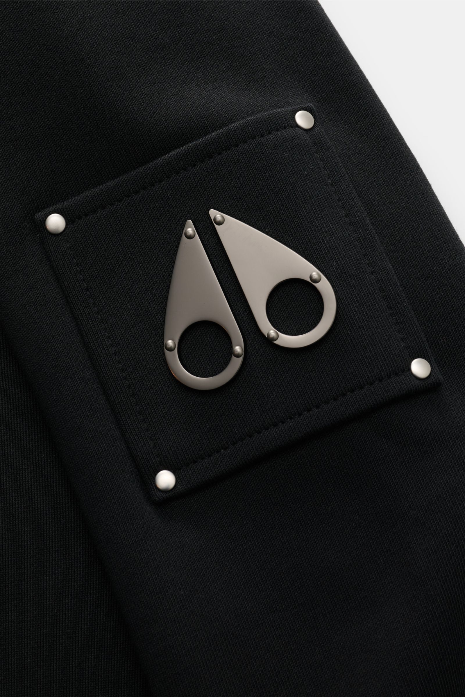 Nahaufnahme des typischen Metall-Logos am Oberarm des Moose Knuckles Sweat-Troyer 'Hartsfield' schwarz, Sweat aus reiner Baumwolle, weicher Griff, Regular Fit, hoch schließender Kragen mit Zipper, elastische Rippbündchen.