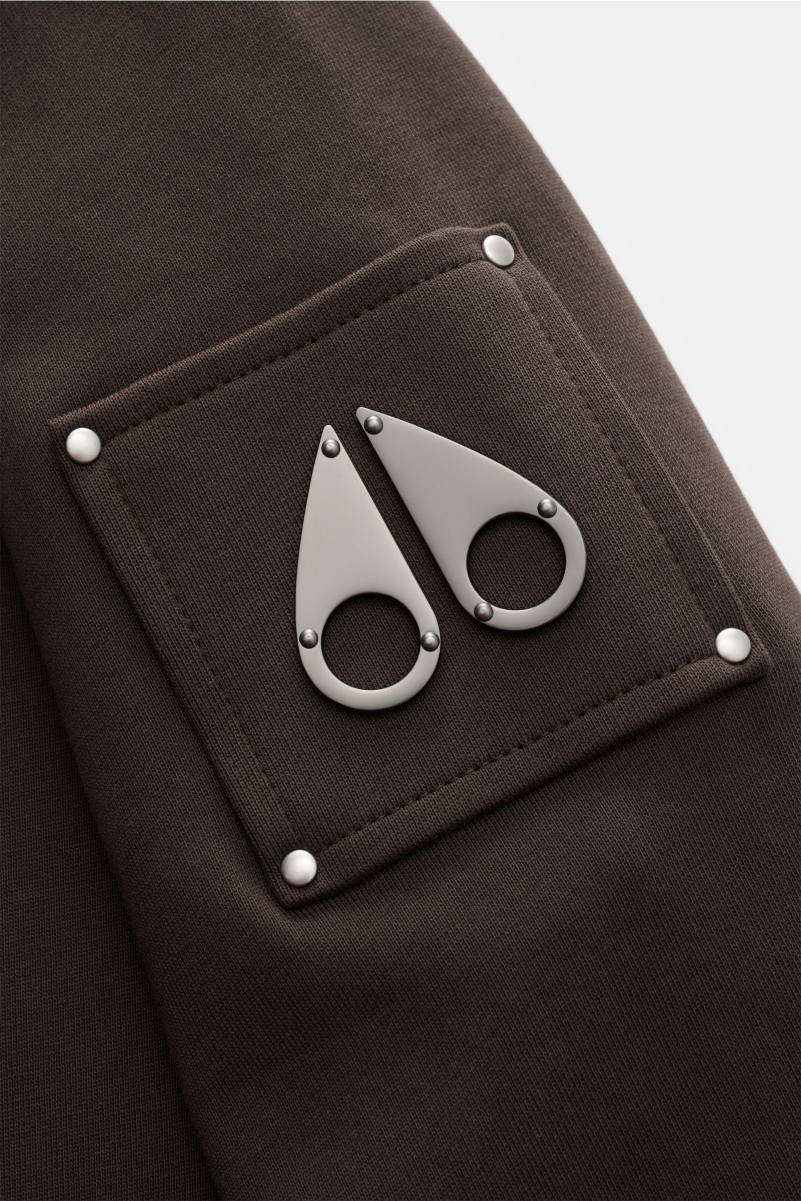 Nahaufnahme des Moose Knuckles Sweat-Troyer 'Hartsfield' graubraun, perspektivisch am Oberarm mit Metall-Logo, reiner Baumwolle, weichem Griff, Regular Fit, hoch schließendem Kragen mit Zipper und elastischen Rippbündchen.