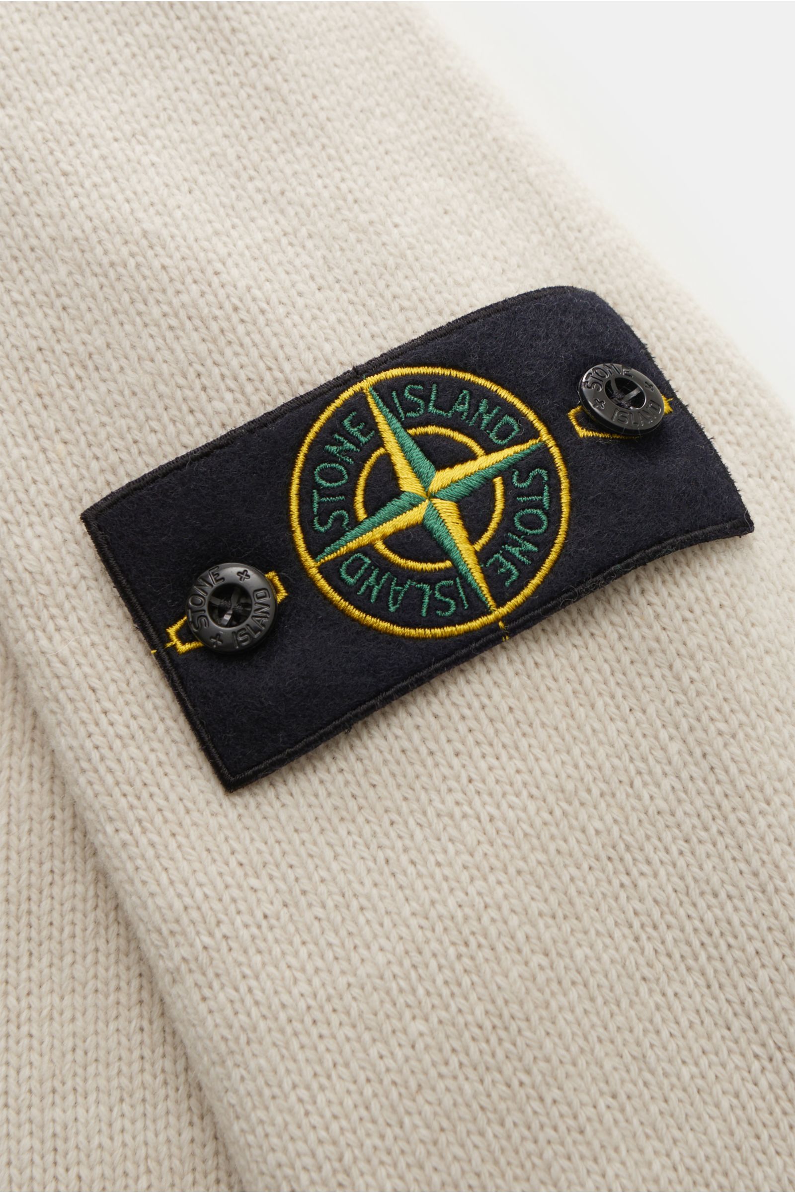 Nahaufnahme der Stone Island Strickjacke 'Lambswool' sand, Fokus auf das schwarze Kompass-Badge mit gelb-grünem Stick und Knöpfen, gestrickt aus weichem Wollmix mit mittelfeinem Strickbild und Filz-Details.