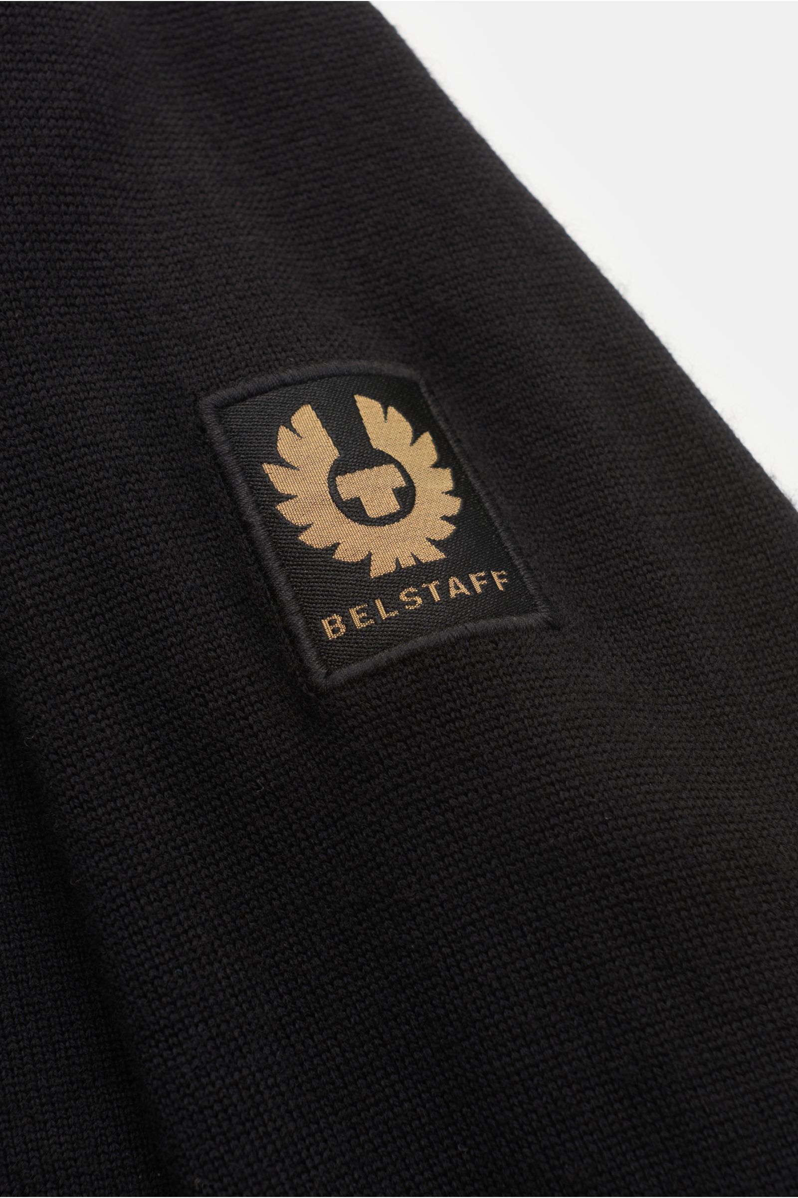 Close-up from a diagonal top perspective of the Belstaff Strickjacke 'Kelby' schwarz sleeve, showing the black knit fabric and the gold and black BELSTAFF logo patch.

Description: Bei BELSTAFF erinnert auch die Strickjacke 'Kelby' an die Wurzeln des L