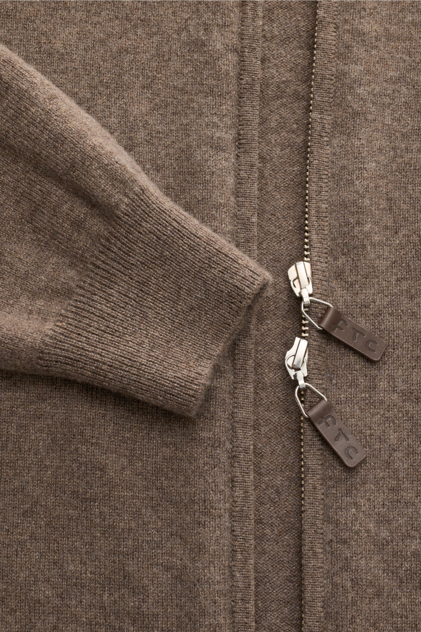 Nahaufnahme der FTC Cashmere Strickjacke graubraun aus Fair-Trade-Cashmere mit Slim Fit, feinem Strickbild, verdecktem Zwei-Wege-Reißverschluss, Leder-Logo-Zipper und geripptem Stehkragen, fotografiert von oben.