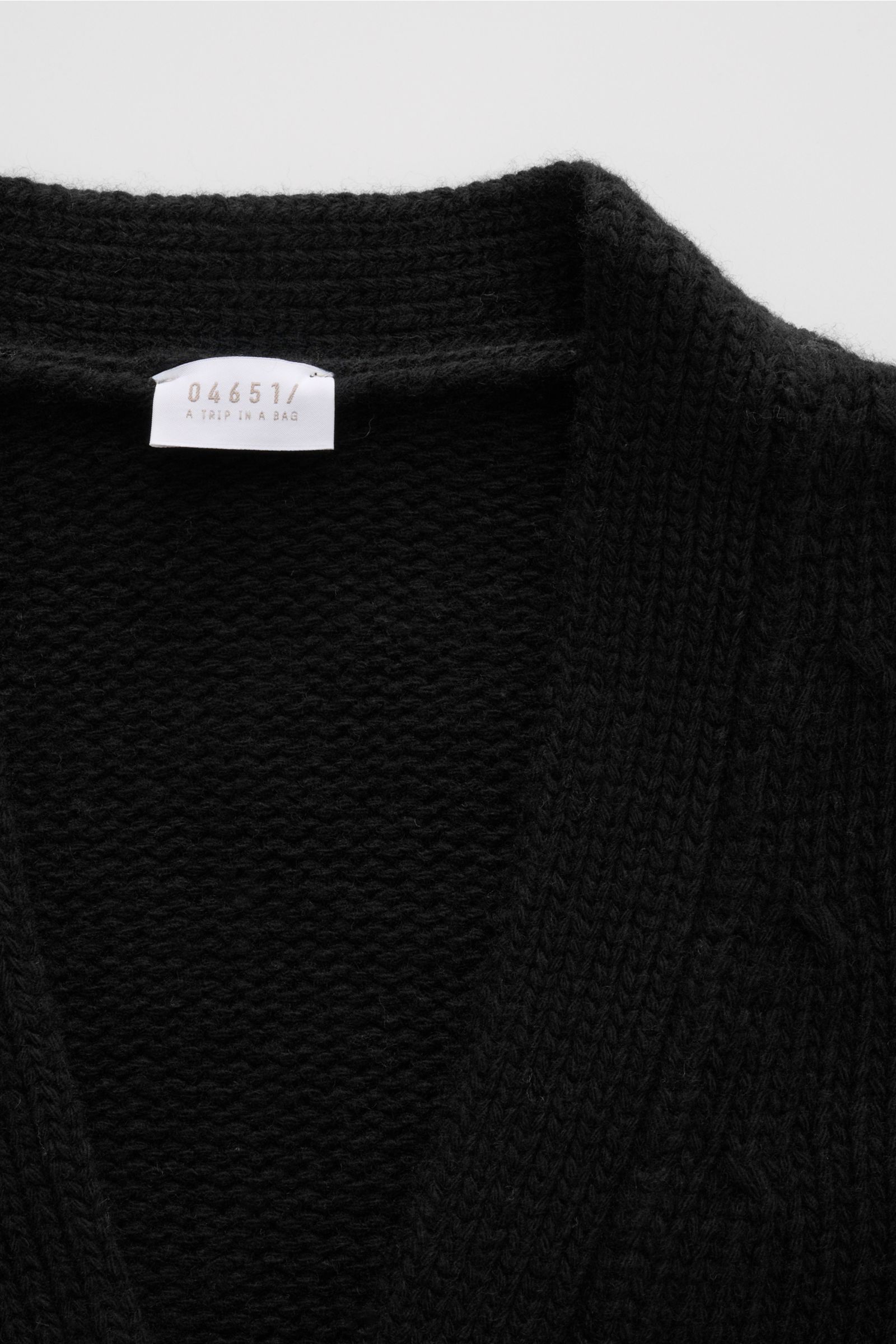 Nahaufnahme des schwarzen 04651/ A trip in a bag Cardigan 'Bold' mit grobem Strickbild, weichem Griff und sichtbarem Label innen am Kragen.