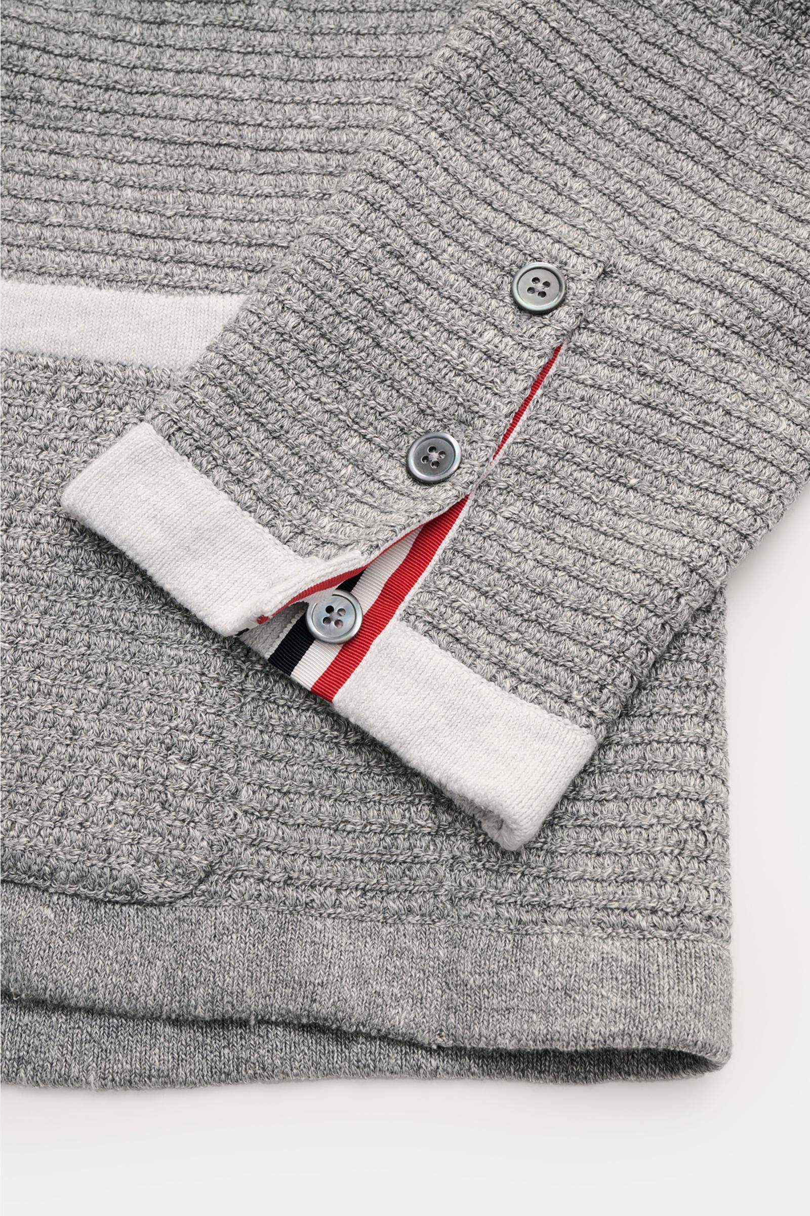 Nahaufnahme des Thom Browne Cardigans hellgrau meliert, Detailansicht des Ärmels mit Tricolore-Ripsband, vier Knöpfen, markantem Strickmuster aus Leinen, Baumwolle und Microfaser.
