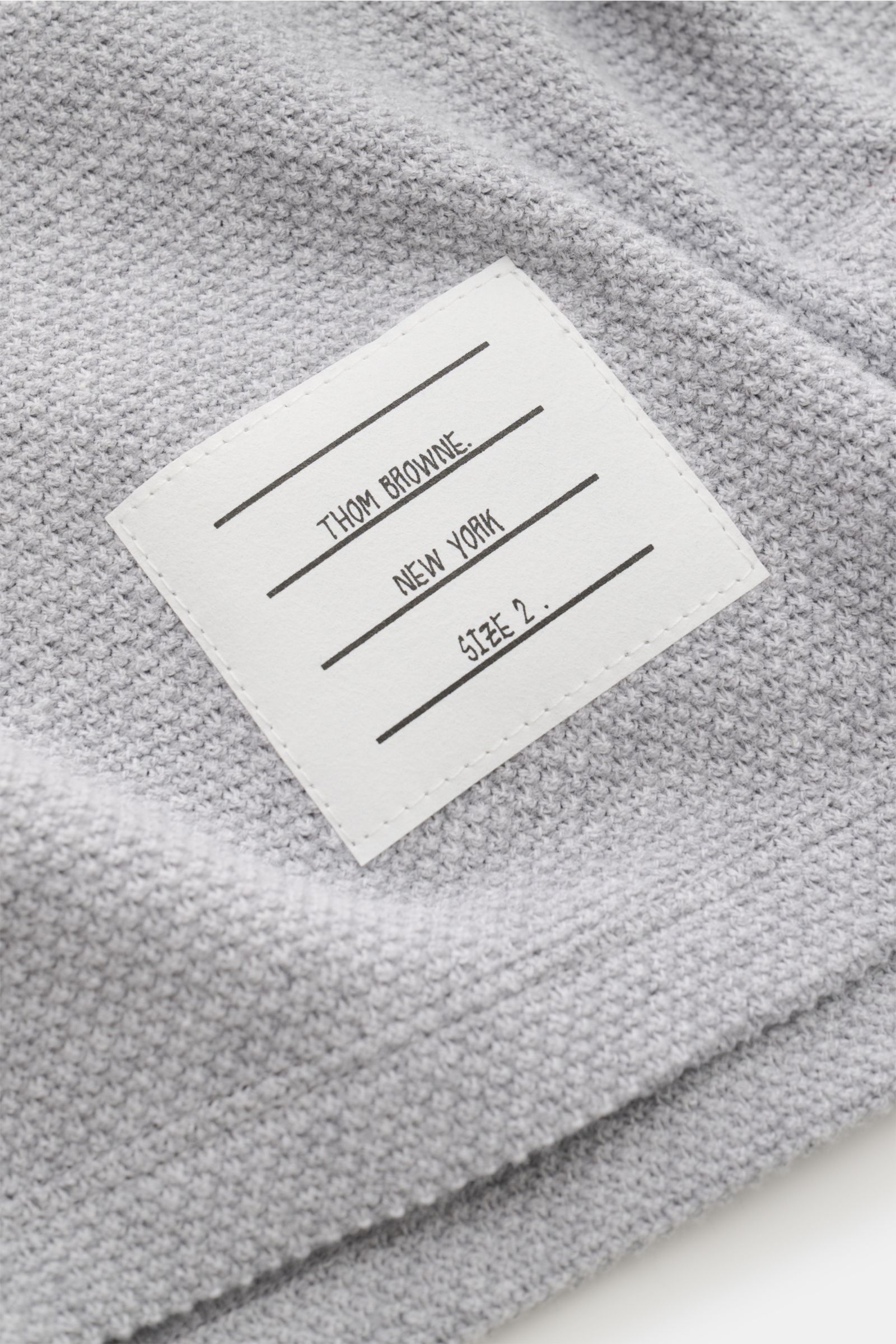 Close-up view of the woven label on the Thom Browne Cardigan light grey mottled, showing finely textured pure cotton knit fabric. This cotton cardigan by THOM BROWNE combines a campus aesthetic with modern accents: distinctive contrasting stripes meet dar