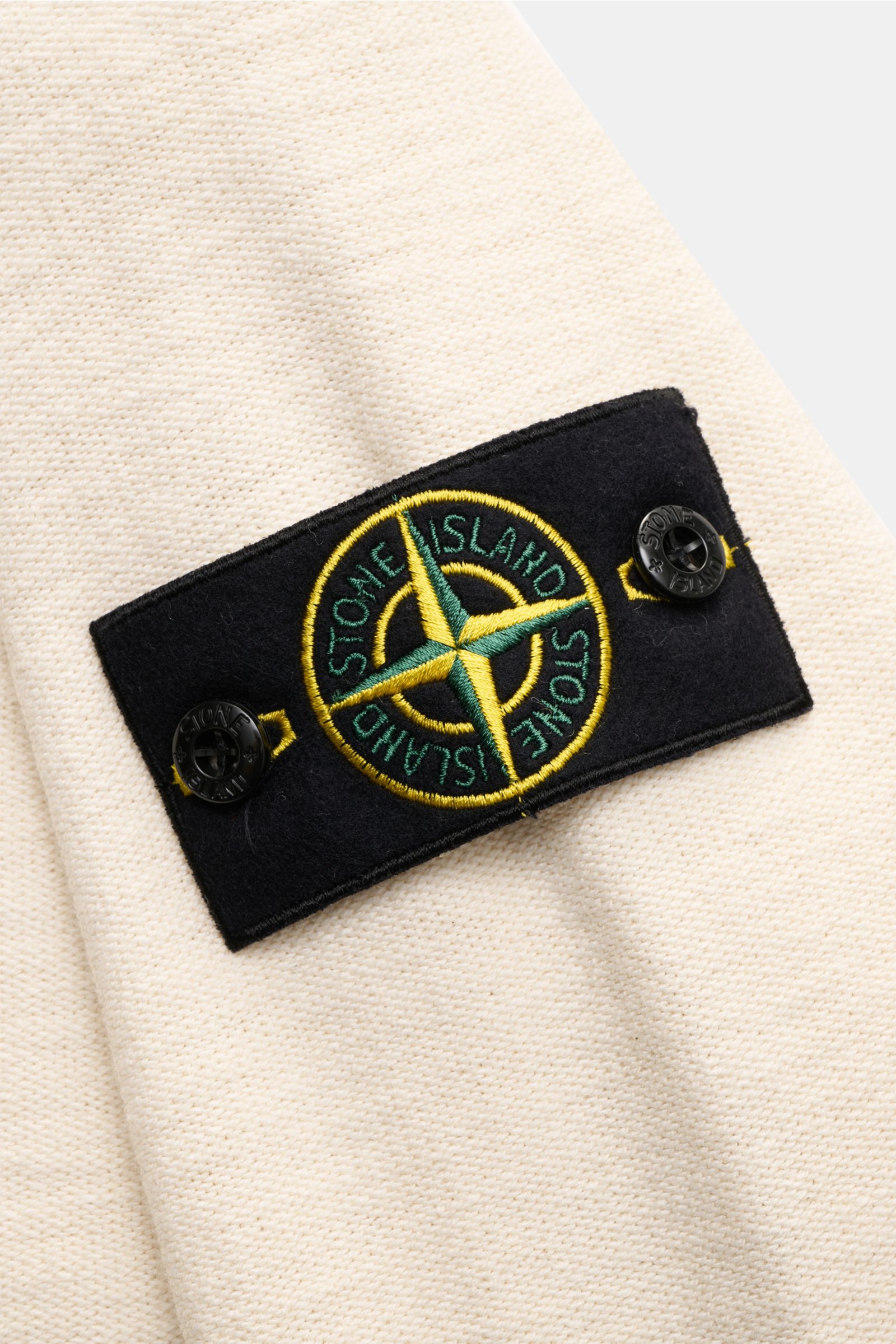Close-up front-side view of the Stone Island Sweatjacke creme showing the black compass badge with yellow and green embroidery on the cream, left-knit cotton-microfiber blend fabric. 

Strick mit Twist: Die sportive Jacke mit Kapuze im modernen Linksst
