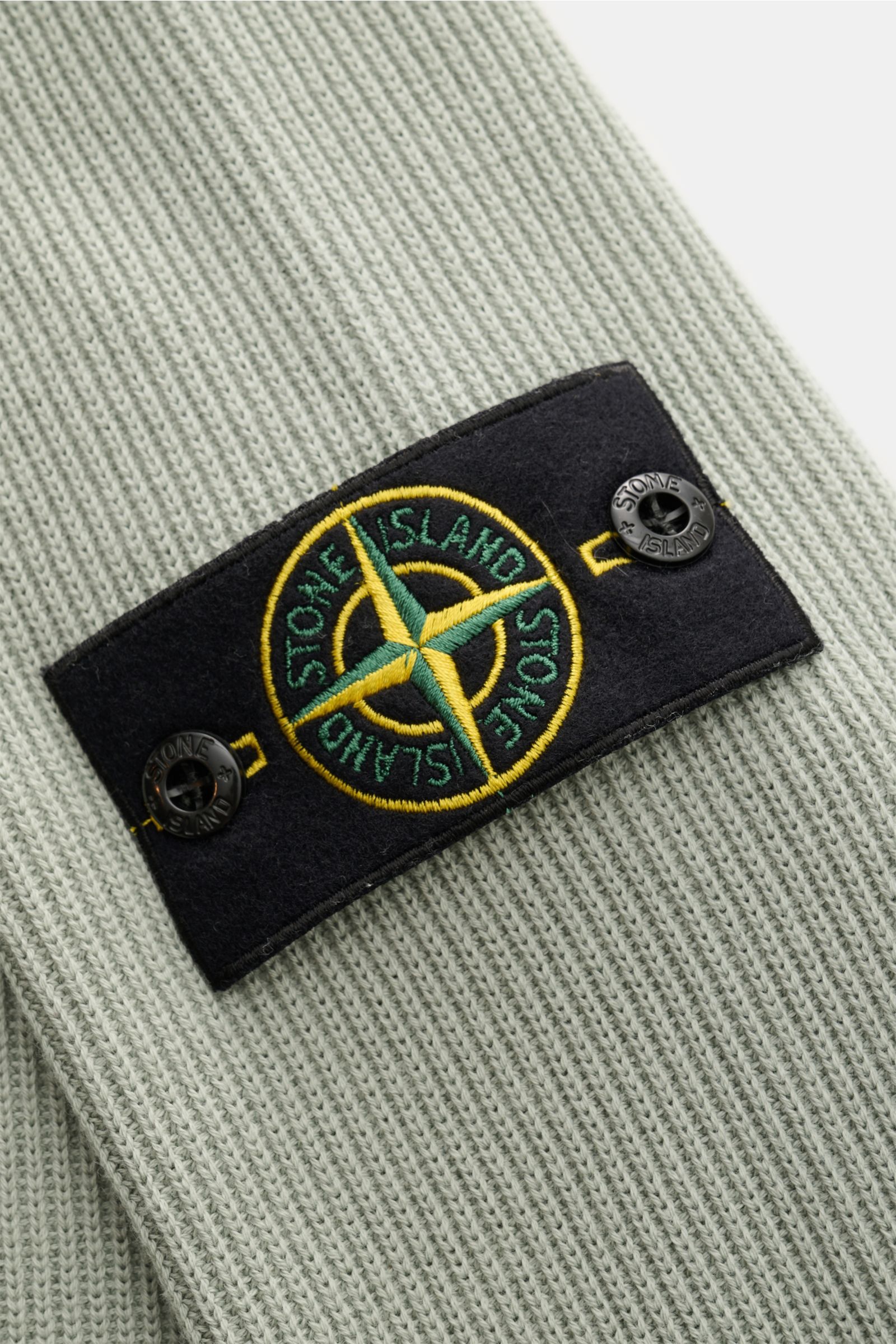 Close-up view of the Stone Island Strickjacke 'Rib' salbei showing the black compass badge with yellow and green embroidery on sage ribbed cotton knit fabric. Zeitloses Layering-Piece: Die Strickjacke von STONE ISLAND wird mit ihrem Rippstrick-Design a