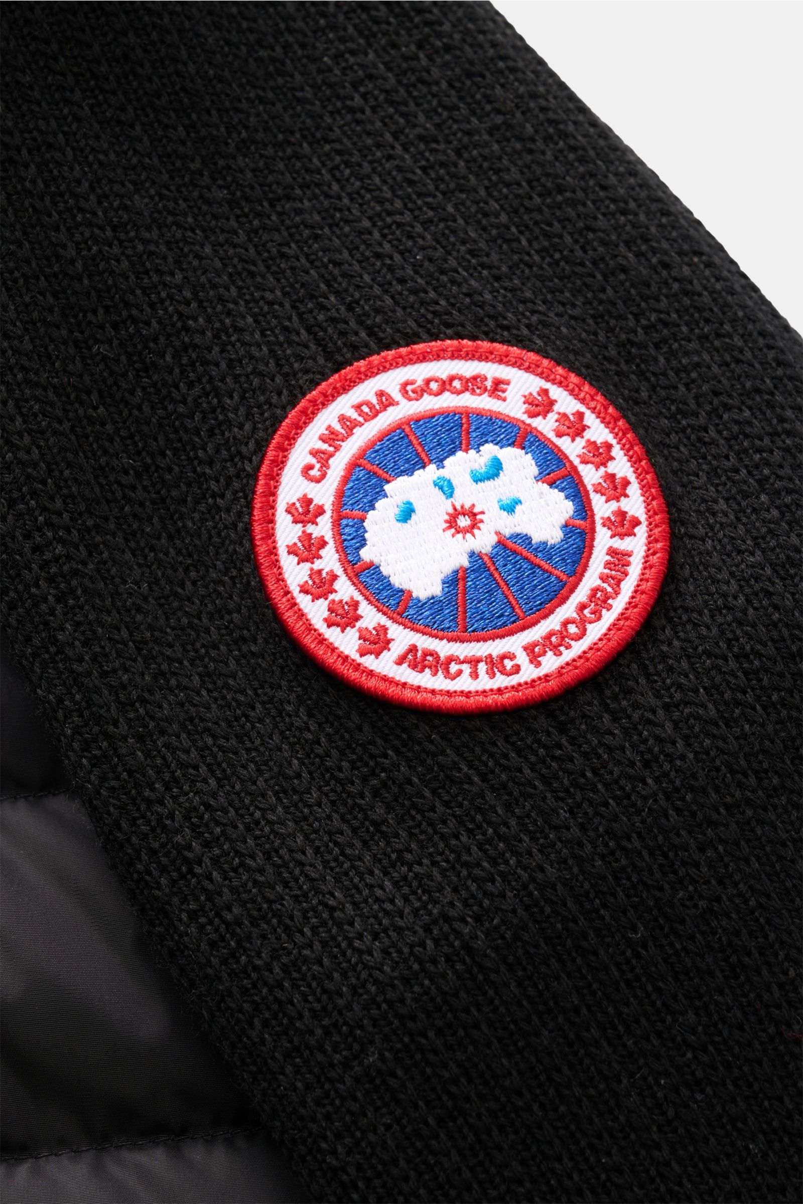 Nahaufnahme aus schräger Perspektive der Canada Goose Strickjacke 'Hybridge Knit' schwarz mit Wollstrick, gesteppter Daunen-Front und rotem Markenlogo am Oberarm.