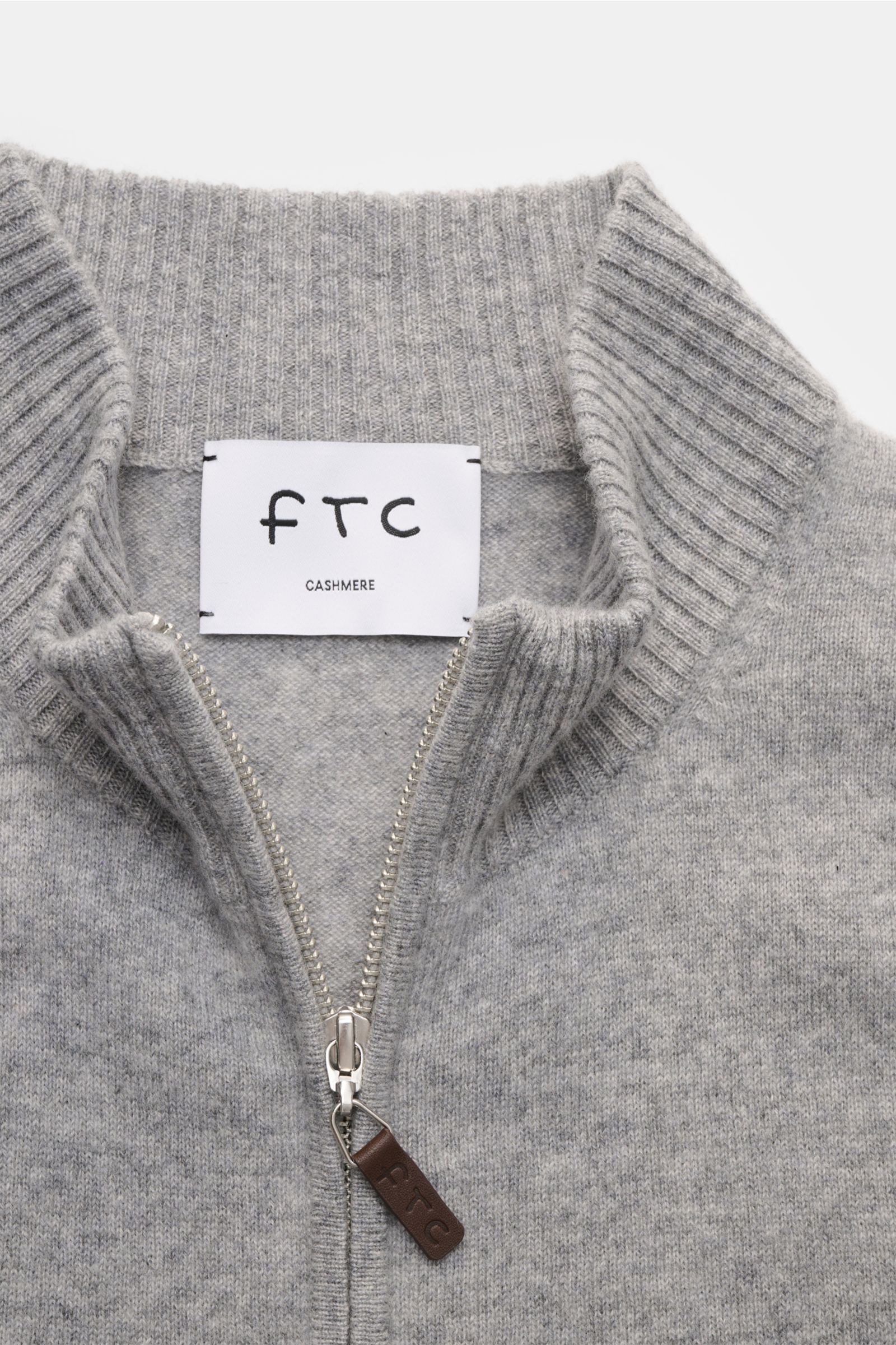 Nahaufnahme der FTC Cashmere Strickjacke grau meliert von oben, zeigt gerippten Stehkragen, feines Strickbild, verdeckten Zwei-Wege-Reißverschluss mit Leder-Logo-Zipper, Slim Fit, OEKO-TEX® zertifiziert, reiner Fair-Trade-Cashmere, sehr weicher Griff, fle