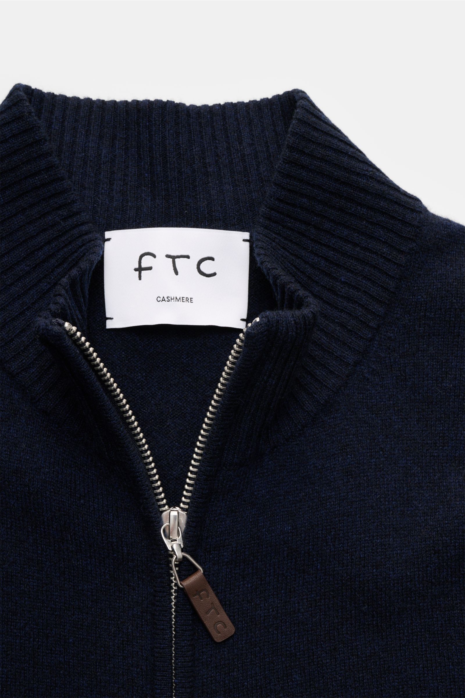 Nahaufnahme der FTC Cashmere Strickjacke navy aus Fair-Trade-Cashmere mit geripptem Stehkragen, verdecktem Zwei-Wege-Reißverschluss und Leder-Logo am Zipper, fein gestrickt und slim fit, fotografiert von oben auf den Kragenbereich.