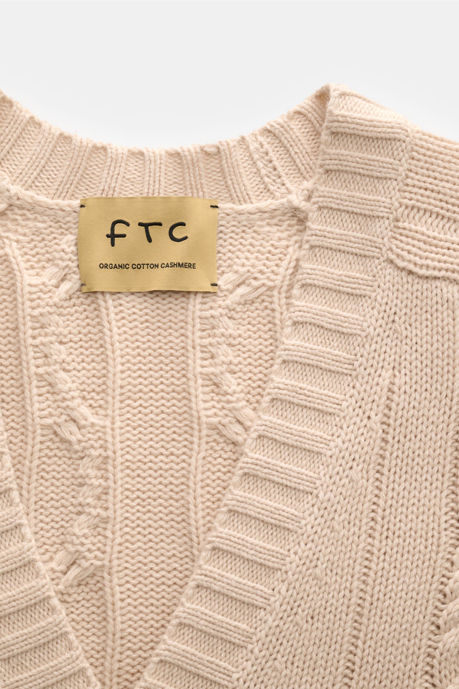 Nahaufnahme des FTC Cardigan beige von oben, zeigt V-Ausschnitt, Zopfstrick-Design, breites Rippbündchen aus Bio-Baumwolle und Fair-Trade-Cashmere.