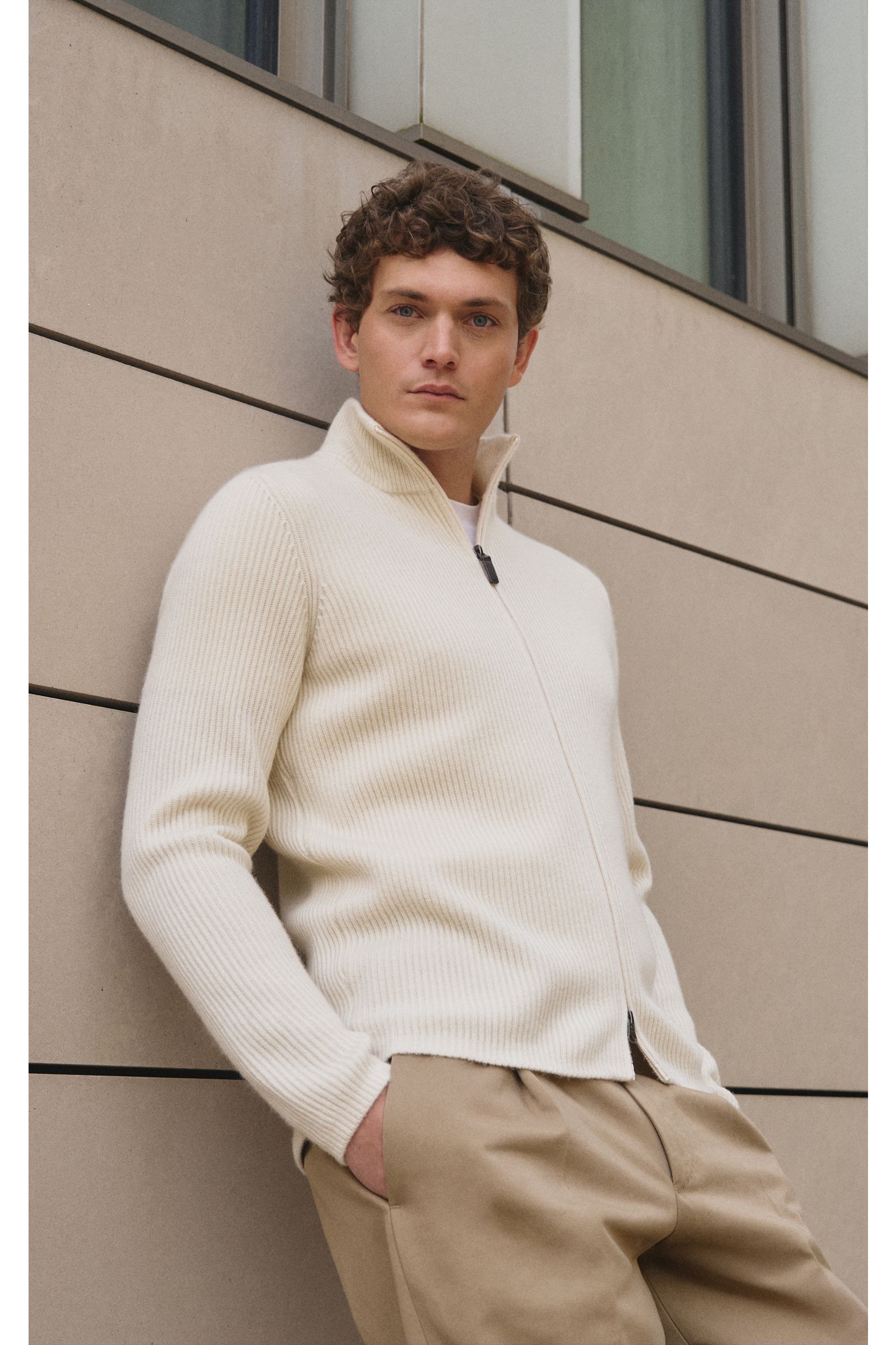 Mann lehnt seitlich an Wand, trägt slim fit von Braun Cashmere Strickjacke offwhite aus mongolischem Cashmere mit Reißverschluss, Patentstrick, hohem Kragen.