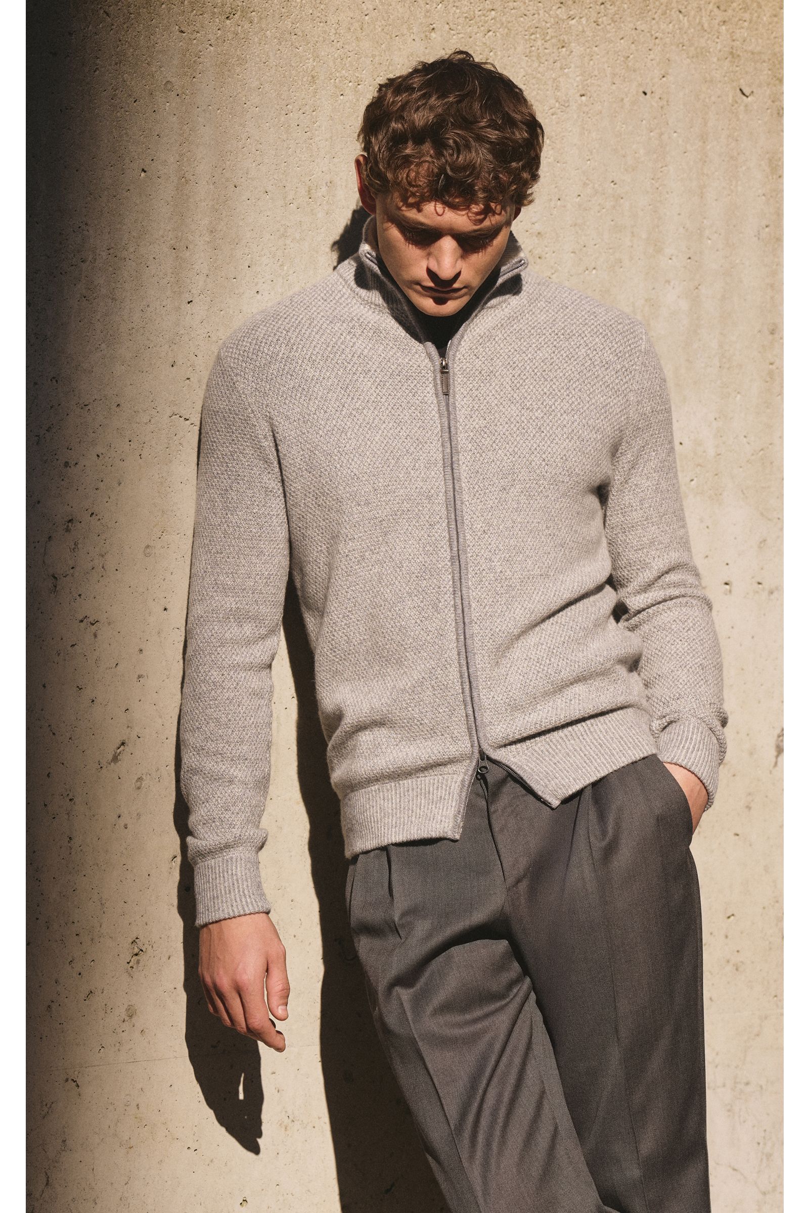 Mann lehnt vor Wand, trägt von Braun Cashmere Strickjacke grau meliert aus mongolischem Cashmere mit Strukturstrick, Slim Fit, Reißverschluss.