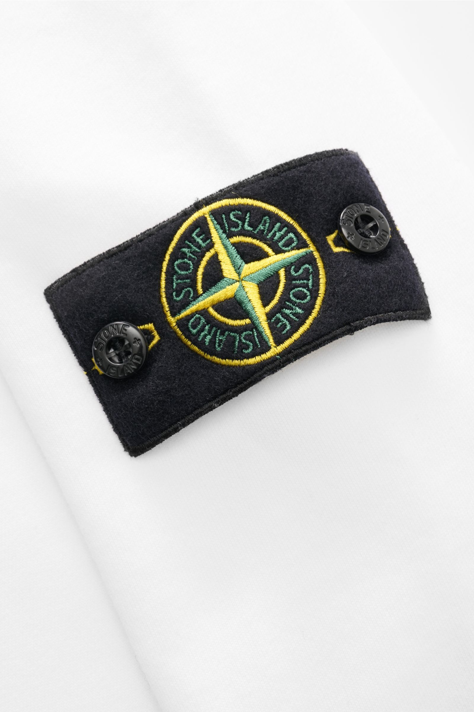 Close-up of the Stone Island Rundhals-Sweatshirt 'Basic Sweat' weiß showing the black compass badge with green and yellow embroidery on white cotton fabric from a top-down angle.

Zeitloses Basic Piece: Das sportive Sweatshirt von STONE ISLAND wird zur