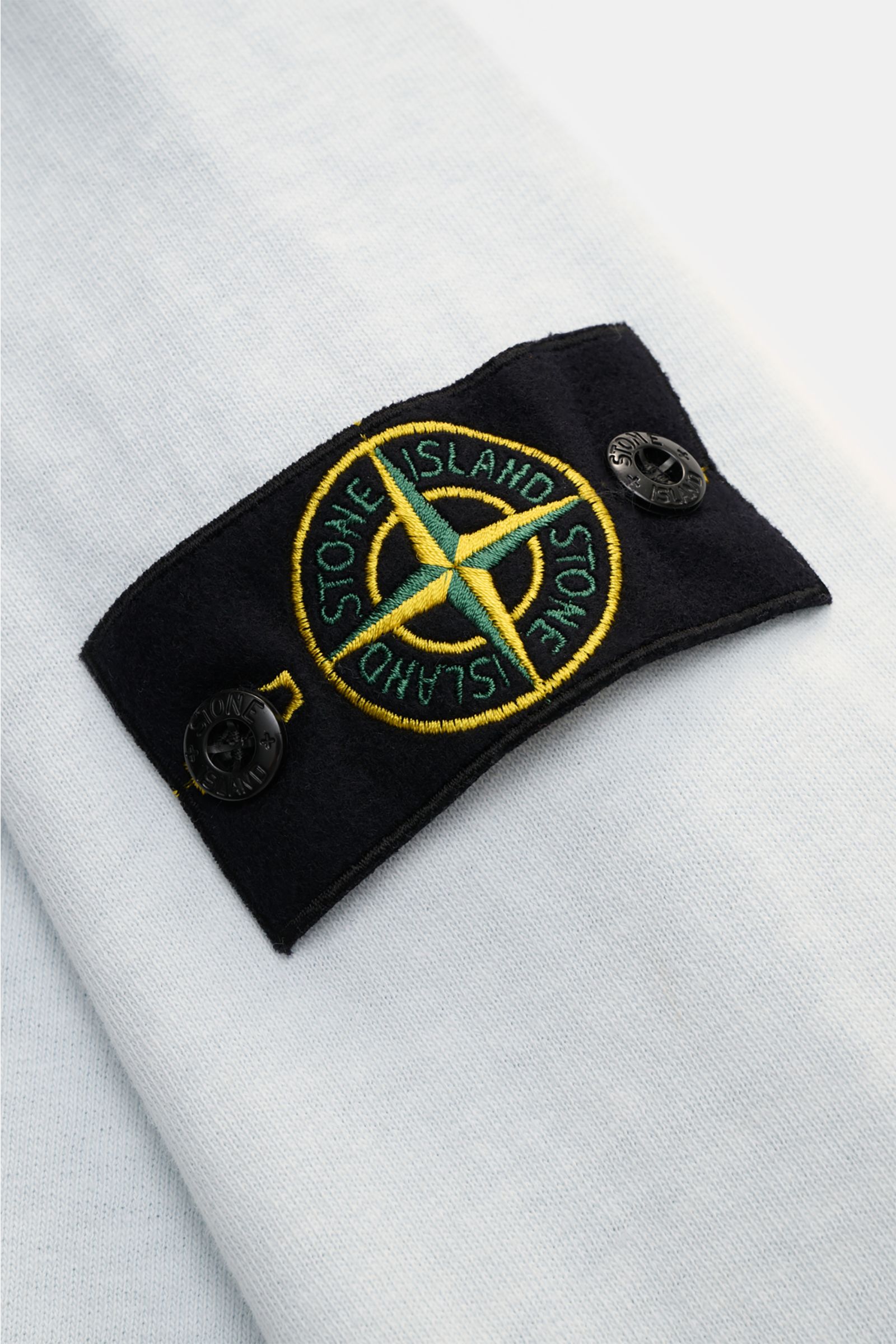 Nahaufnahme des Stone Island Rundhals-Sweatshirt 'Washed Sweat' pastellblau, Fokus auf das Kompass-Badge am Ärmel mit weichem Baumwollstoff und washed Effekt.