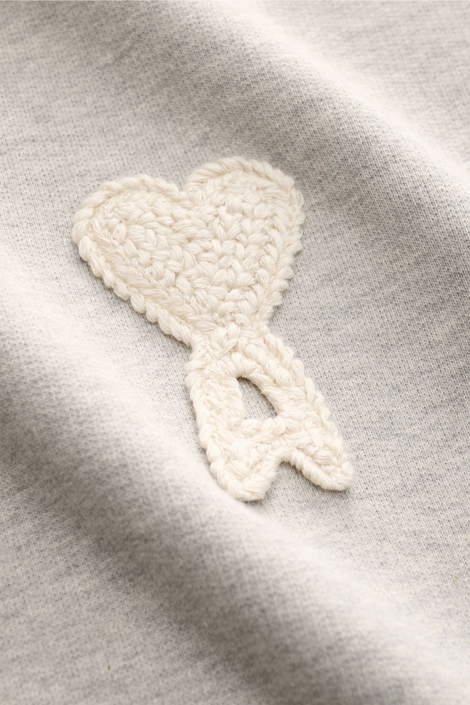 Nahaufnahme des markanten Herz-Stitchings auf dem Ami Paris Sweatshirt 'Crewneck' grau meliert aus leicht elastischer Baumwolle, Slim Fit mit Rundhalsausschnitt und flexiblen Rippbündchen, fotografiert aus schräger Frontperspektive.