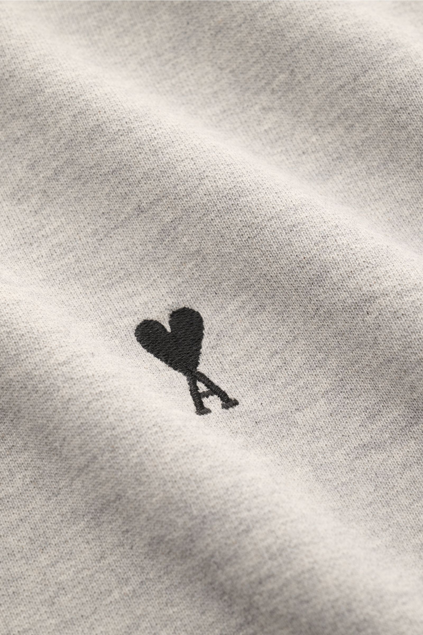 Close-up front view of the Ami Paris Sweatshirt hellgrau meliert showing the subtle black heart-stitching on light gray cotton fabric. Ein dezentes Herz-Stitching auf der Brust verleiht diesem Baumwoll-Sweatshirt von AMI PARIS seine besondere Note und 