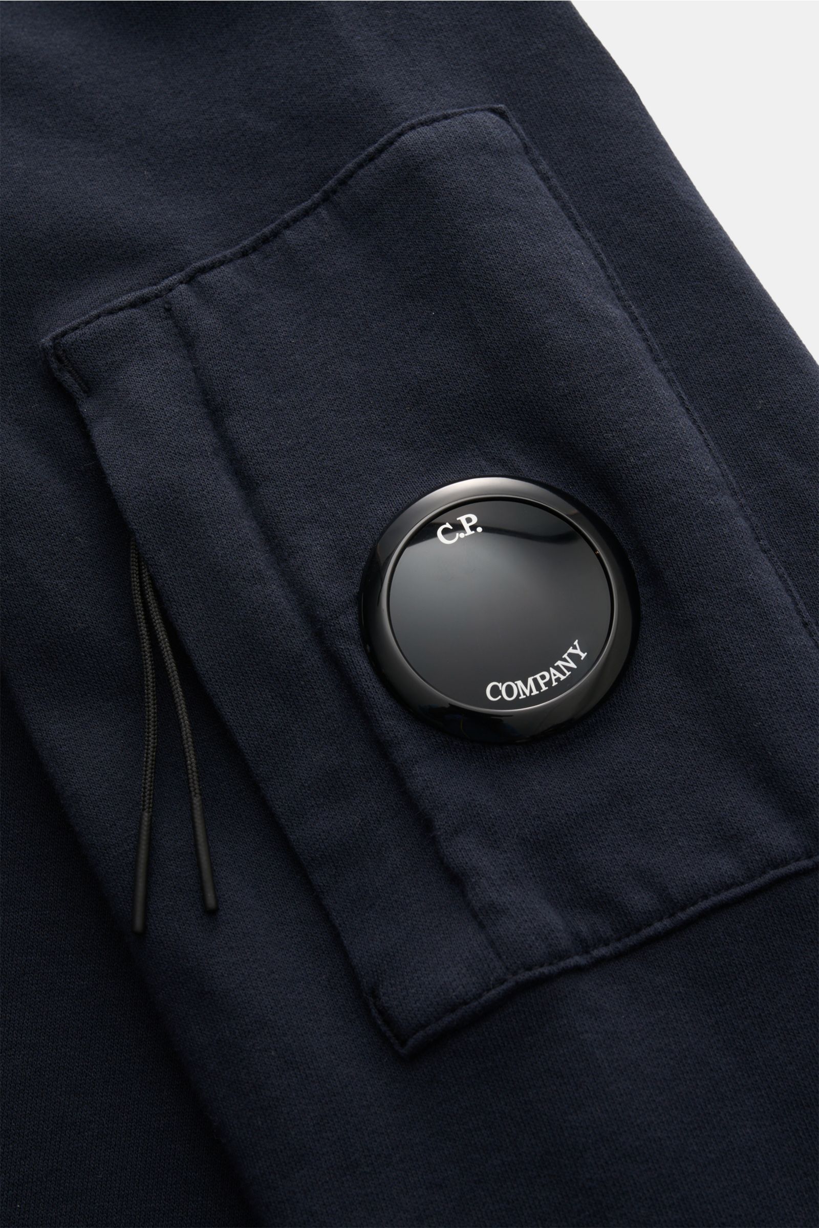 Close-up view of the navy C.P. Company Sweatshirt showing the garment-dyed cotton fabric, slim fit sleeve with a zipper pocket, and the iconic black lens detail with 'C.P. Company' text on the upper arm. The image highlights the soft texture and flexible 