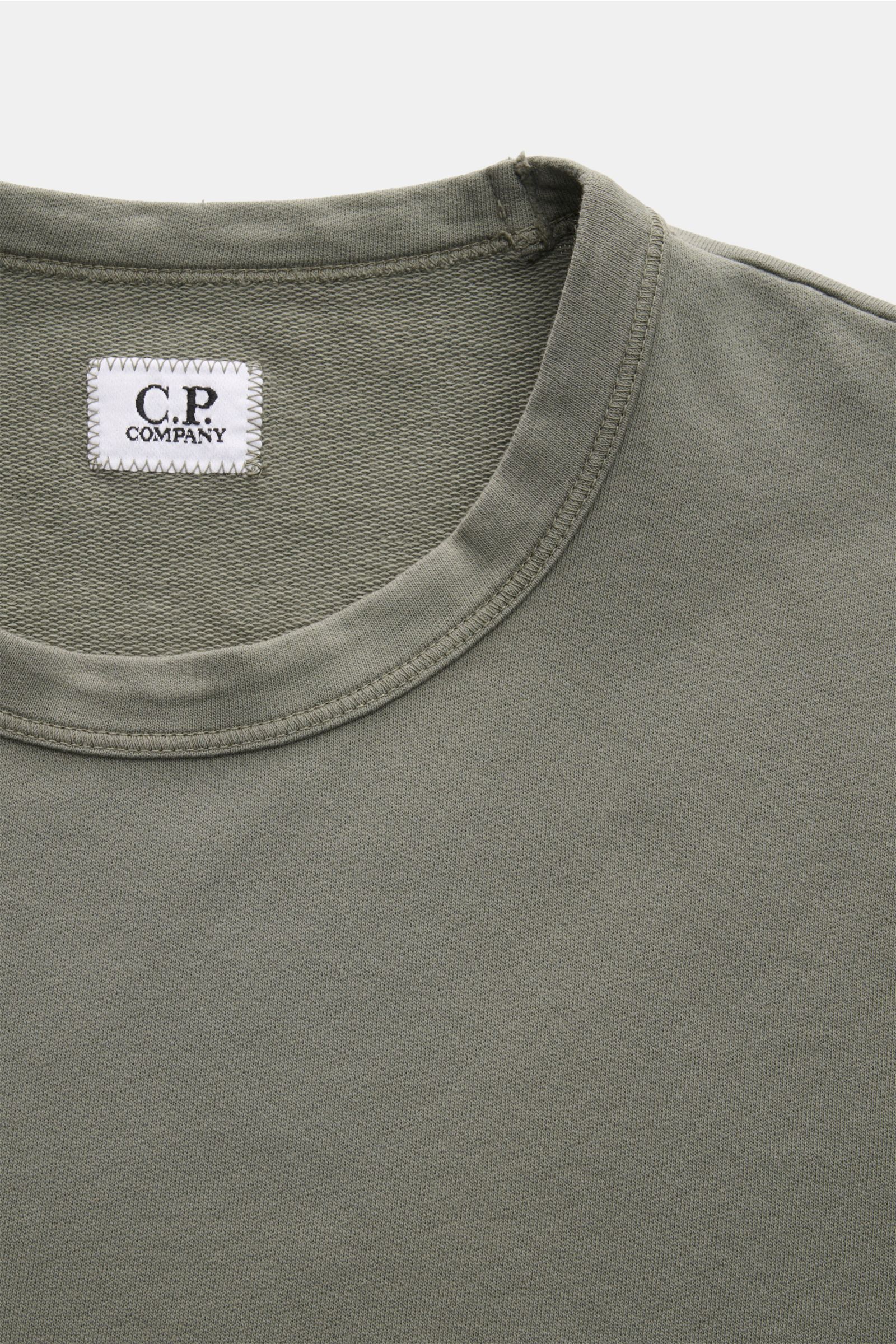Close-up front top view of a C.P. Company Sweatshirt salbei in a slim fit, garment-dyed light olive green cotton with a round neckline and visible white label, showcasing soft fabric texture and neat stitching. 