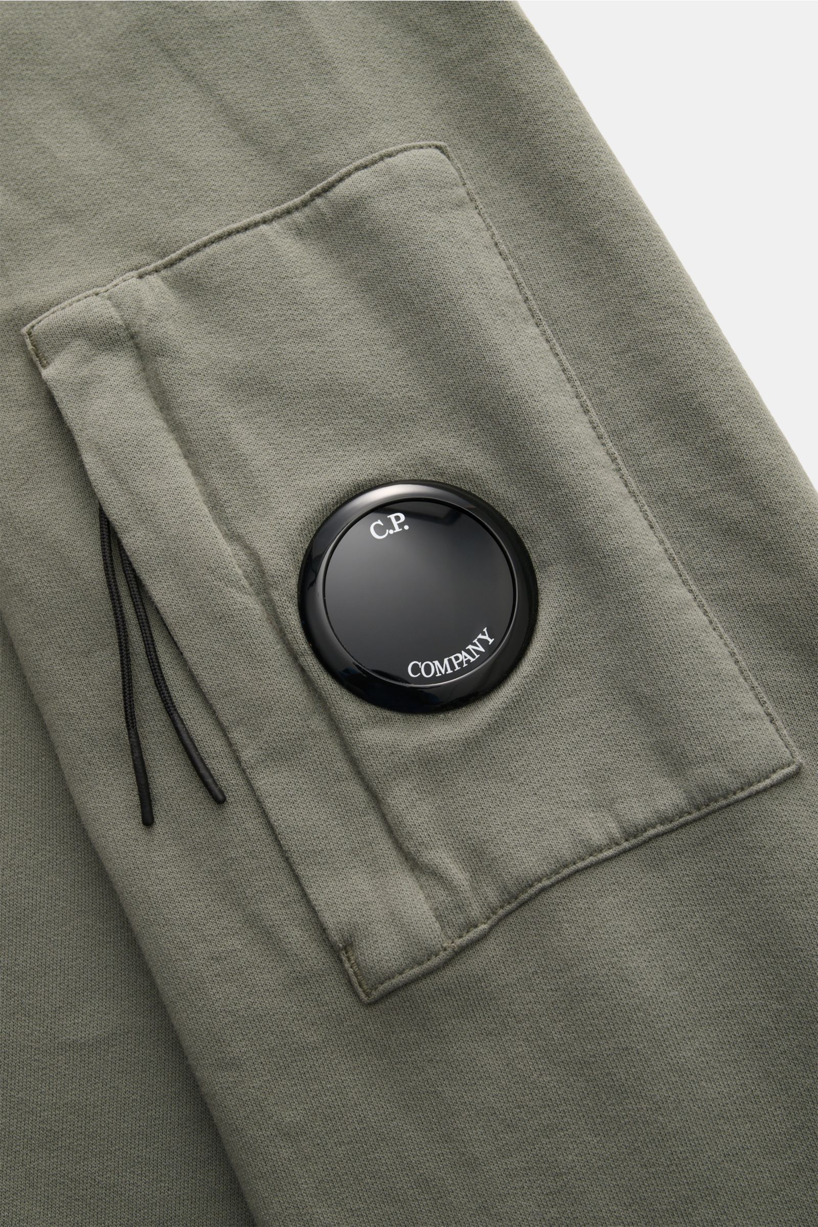 Close-up view of the C.P. Company Sweatshirt salbei sleeve showing the garment-dyed soft cotton fabric, slim fit, round neck, upper arm zipper pocket, and iconic black lens detail. 