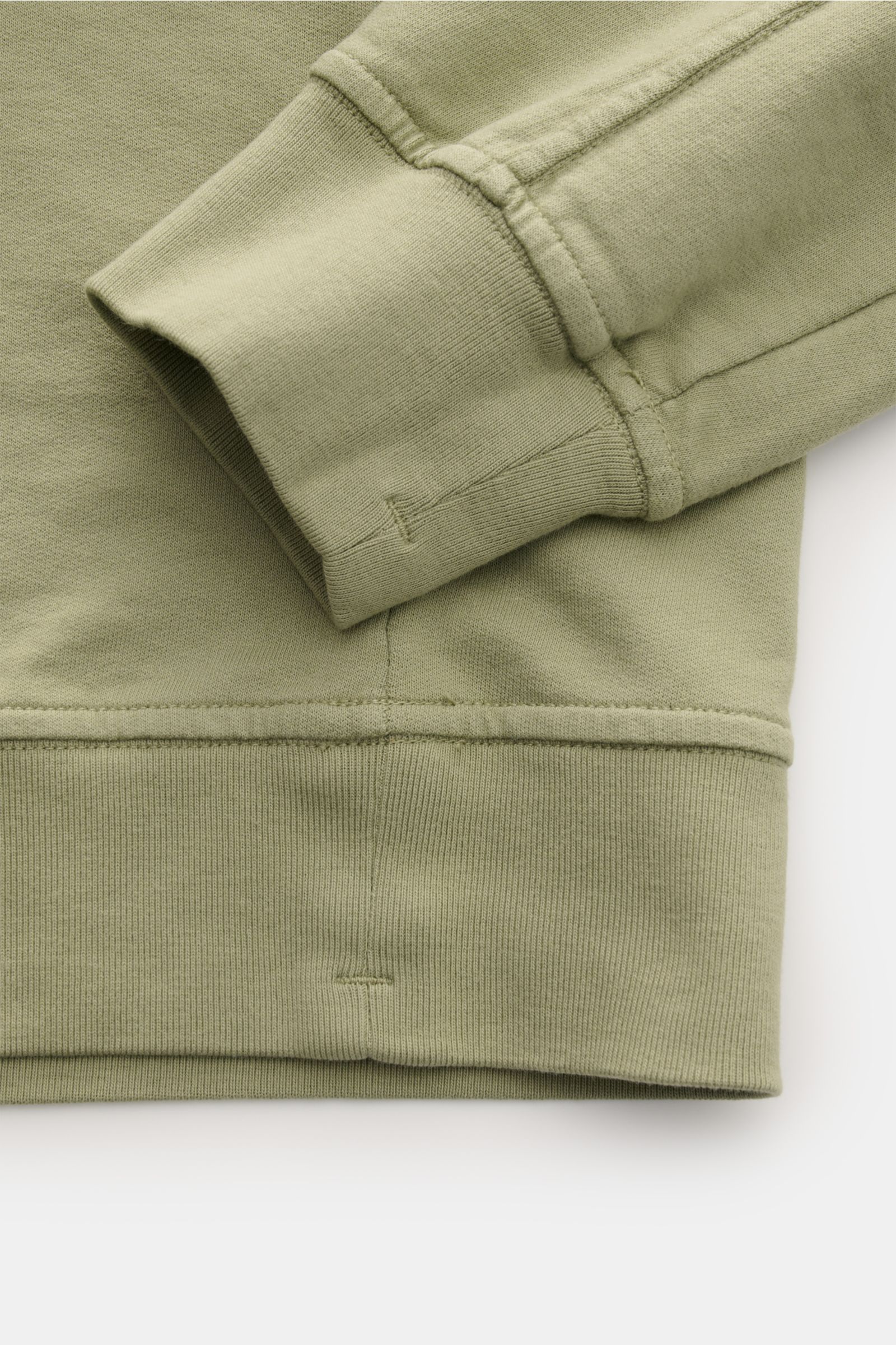 Close-up of the cuff and waistband of the C.P. Company Sweatshirt oliv, showing garment-dyed olive cotton fabric from a side angle, highlighting ribbed flexible cuffs and hem with detailed stitching and lens detail on the sleeve pocket. 